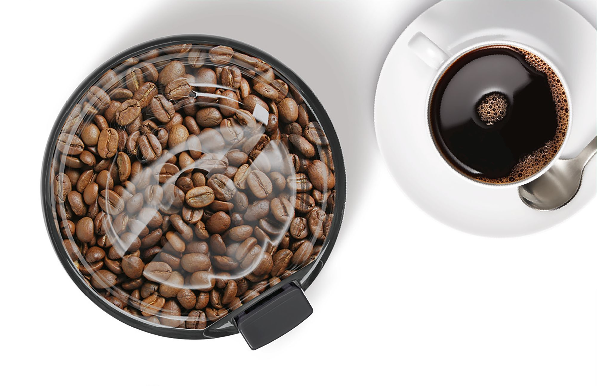 Coffee beans in a grinder next to a cup of coffee on a white surface.