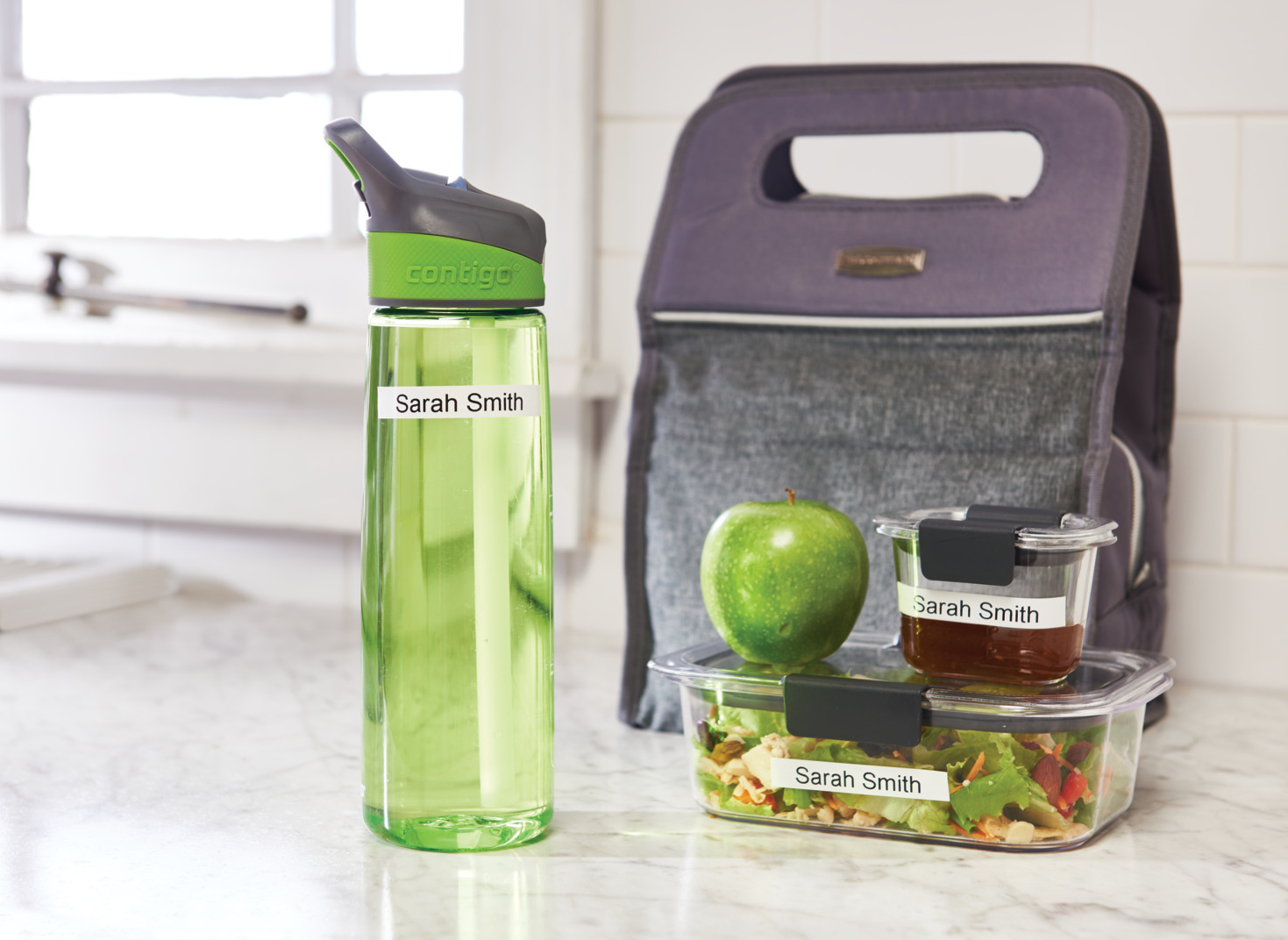 Eine Mittagessenszene mit einer Wasserflasche, einer Lunchtasche, einem grünen Apfel und beschrifteten Behältern.