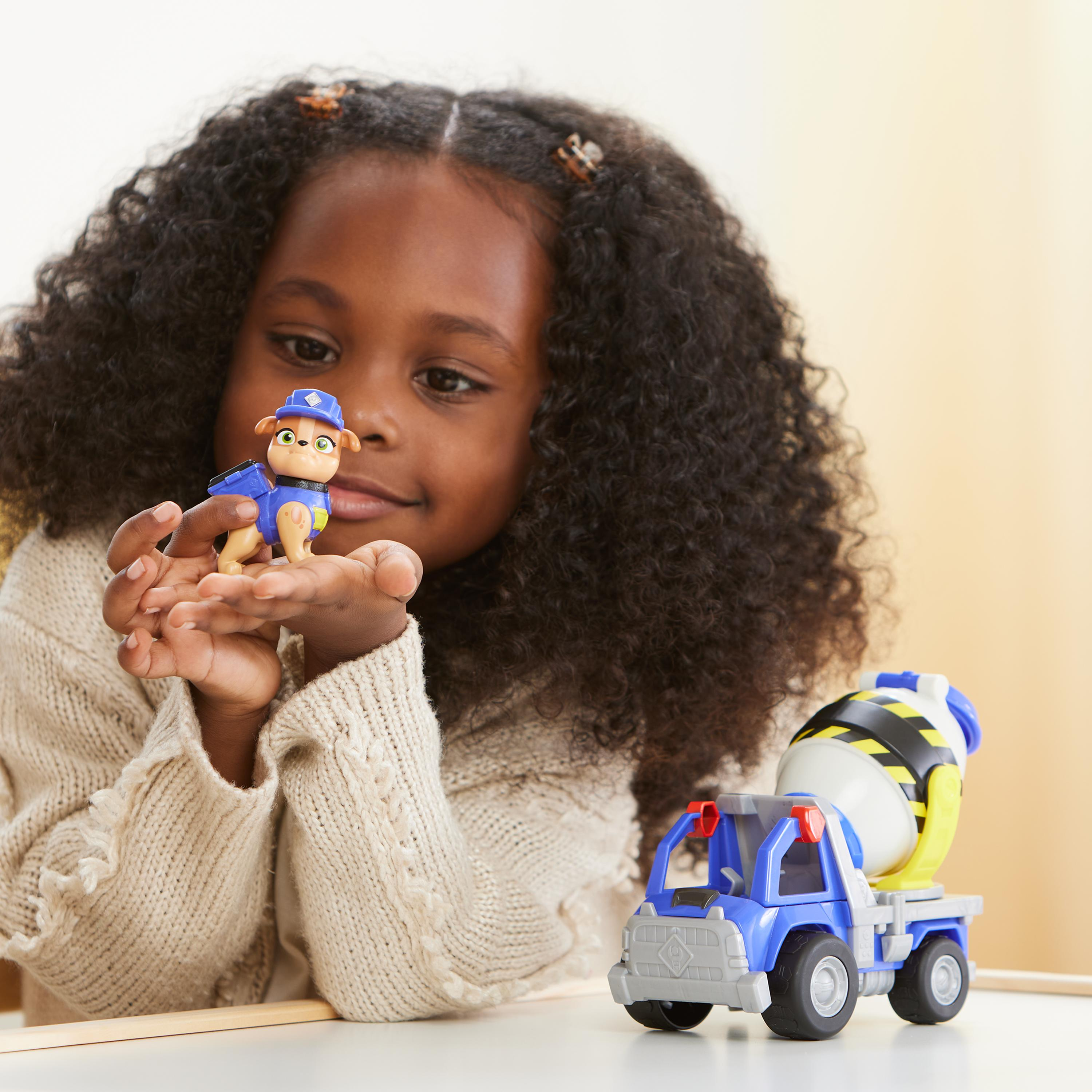 Mädchen betrachtet Paw Patrol-Figur in der Hand. Blau-gelber Spielzeug-LKW rechts.