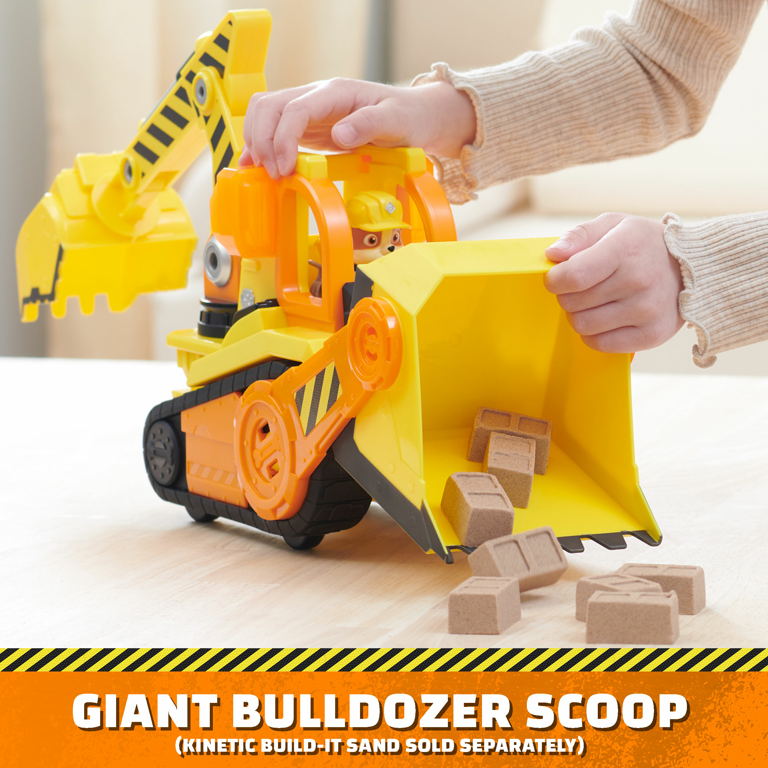 Ein Kind spielt mit einem gelben Bulldozerspielzeug und schiebt braune Blöcke in die Schaufel. Text: Giant Bulldozer Scoop.