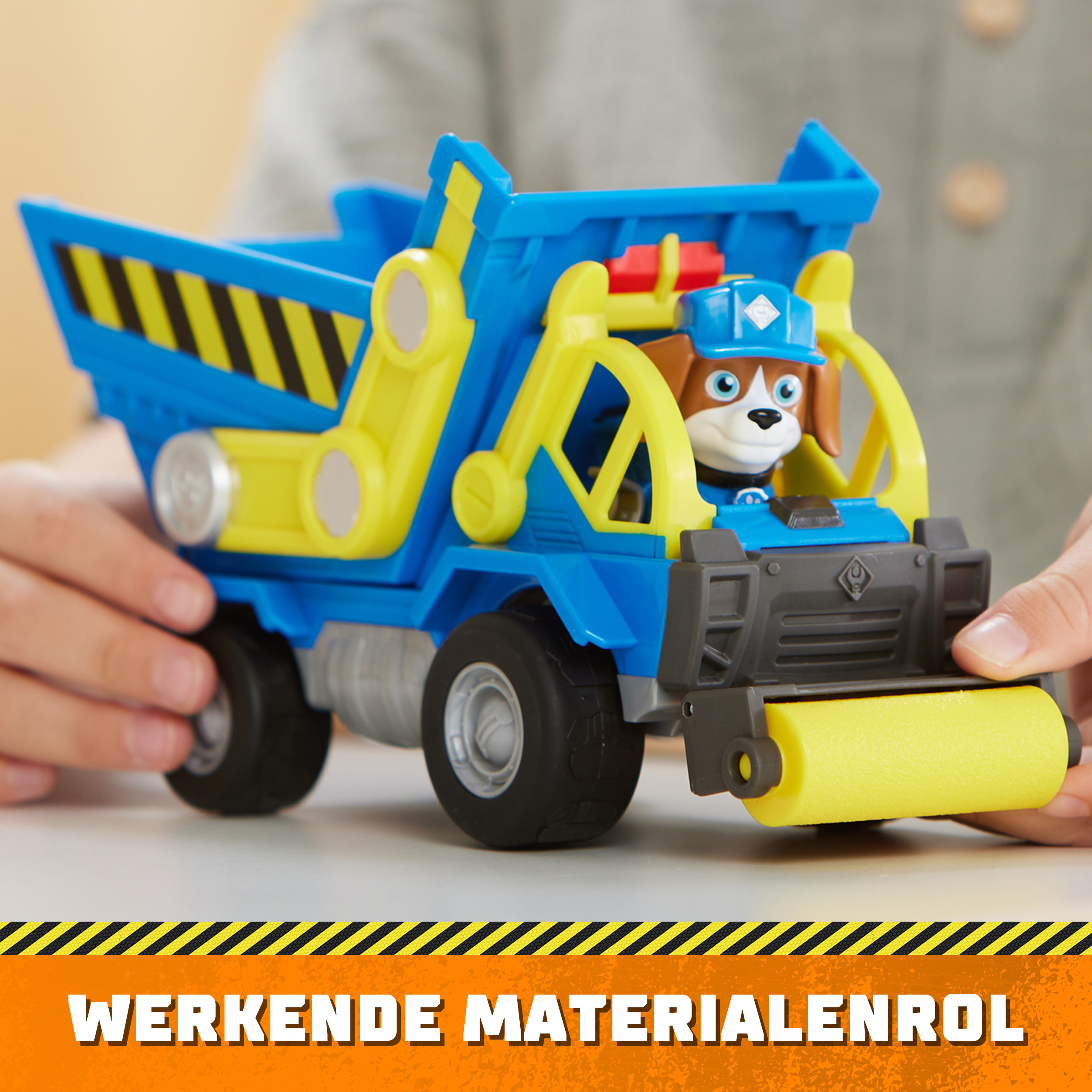 Ein blau-gelber Spielzeug-LKW. Eine Hundefigur sitzt am Steuer. Text: "WERKENDE MATERIALENROL".