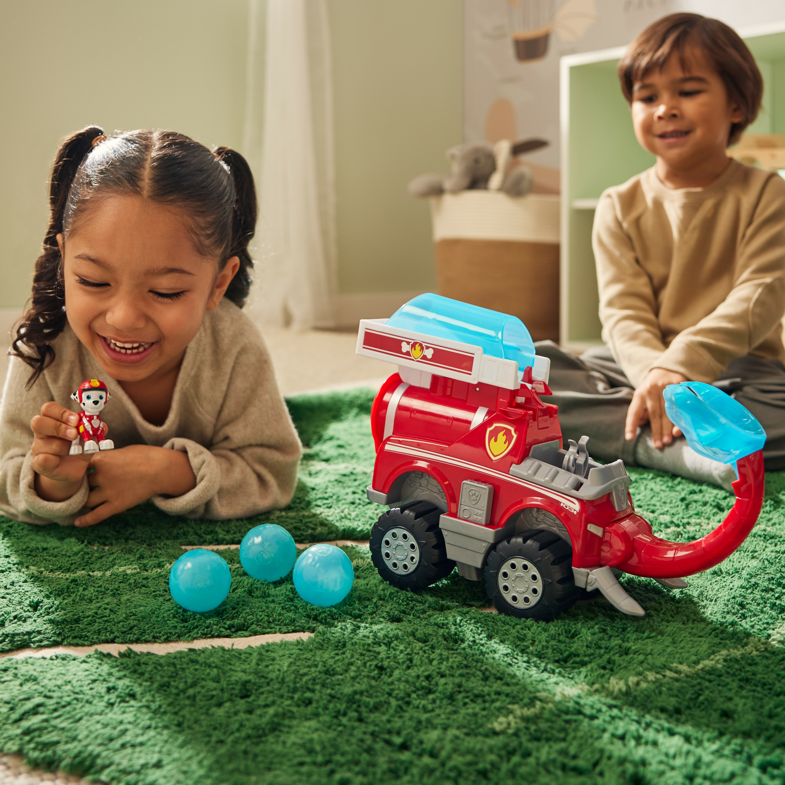 Zwei Kinder spielen mit einem roten Feuerwehrauto und Bällen. Ein Mädchen hält eine Spielfigur, ein Junge lächelt.
