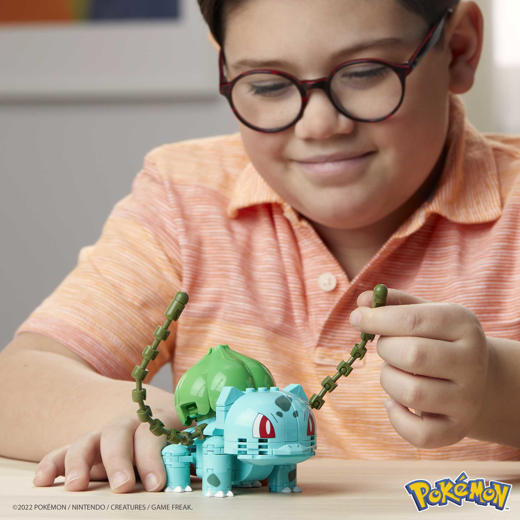 Ein Junge mit Brille spielt mit einem Bulbasaur-Spielzeug. Das Spielzeug ist blau und grün.