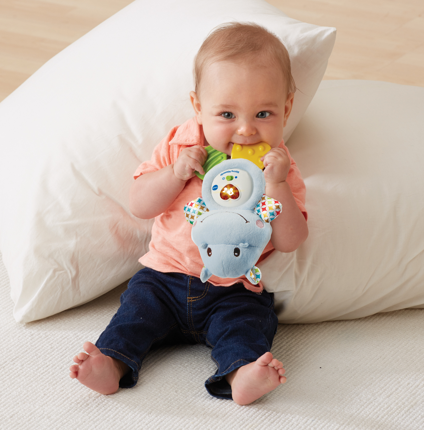 Ein Baby sitzt mit einem Spielzeug im Mund, dahinter ein Kissen.
