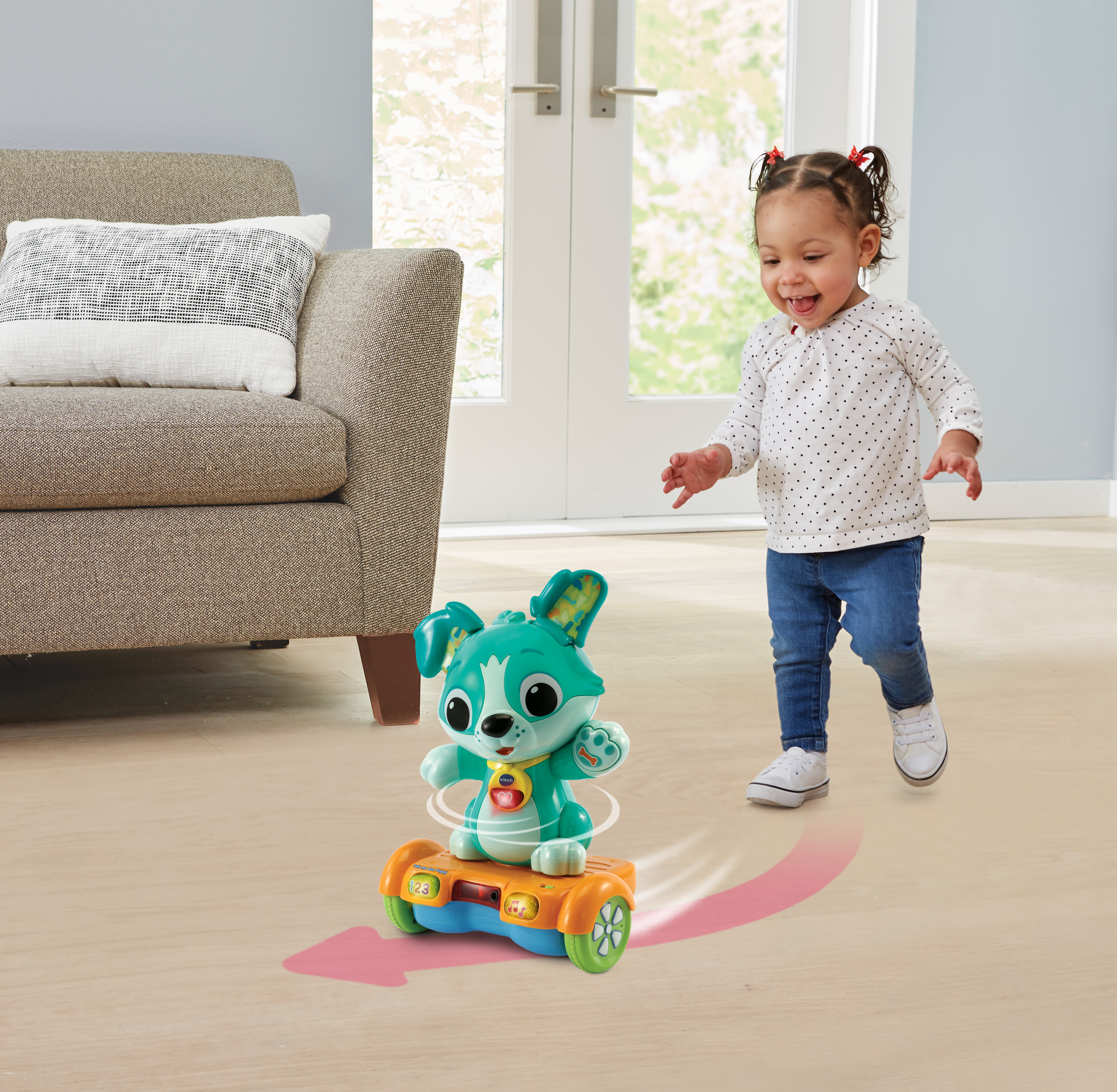 Ein Kleinkind lächelt und greift nach einem Spielzeughund auf einem Hoverboard. Drinnen, Holzboden, beiges Sofa.