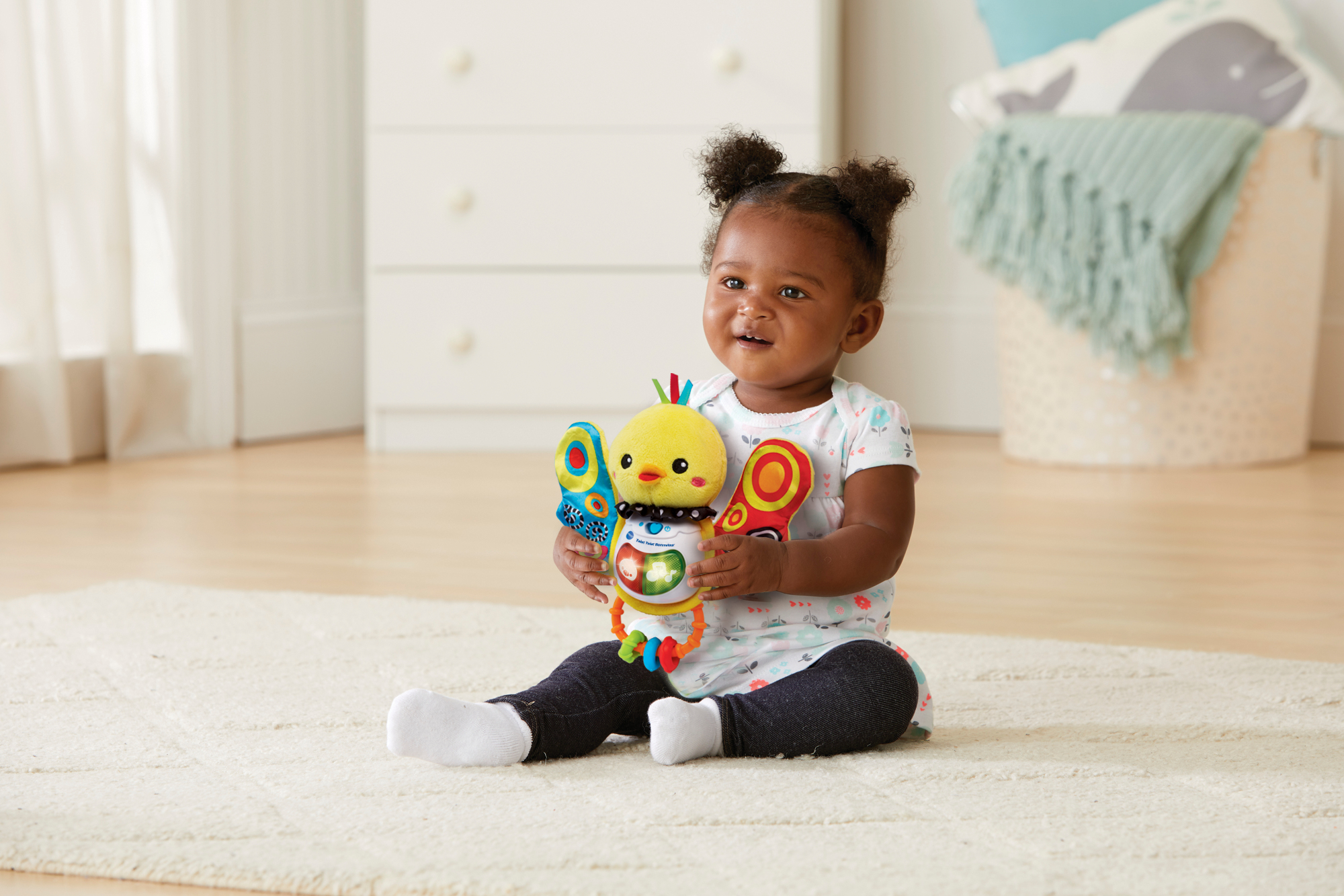 Niemowlę siedzi na dywanie, trzymając kolorową zabawkę. Komoda i kosz w tle.