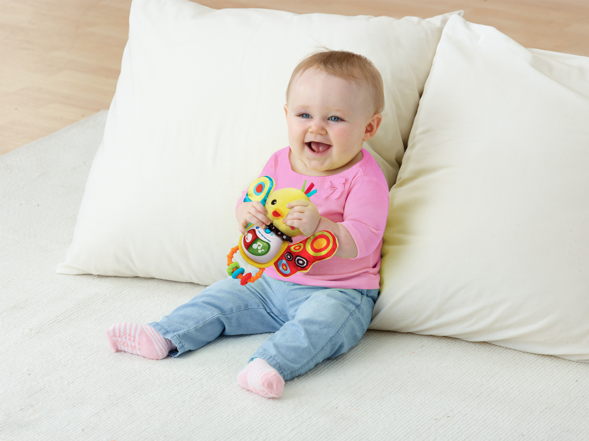 Niemowlę siedzi uśmiechnięte, trzymając kolorową zabawkę, z dużymi poduszkami w tle.