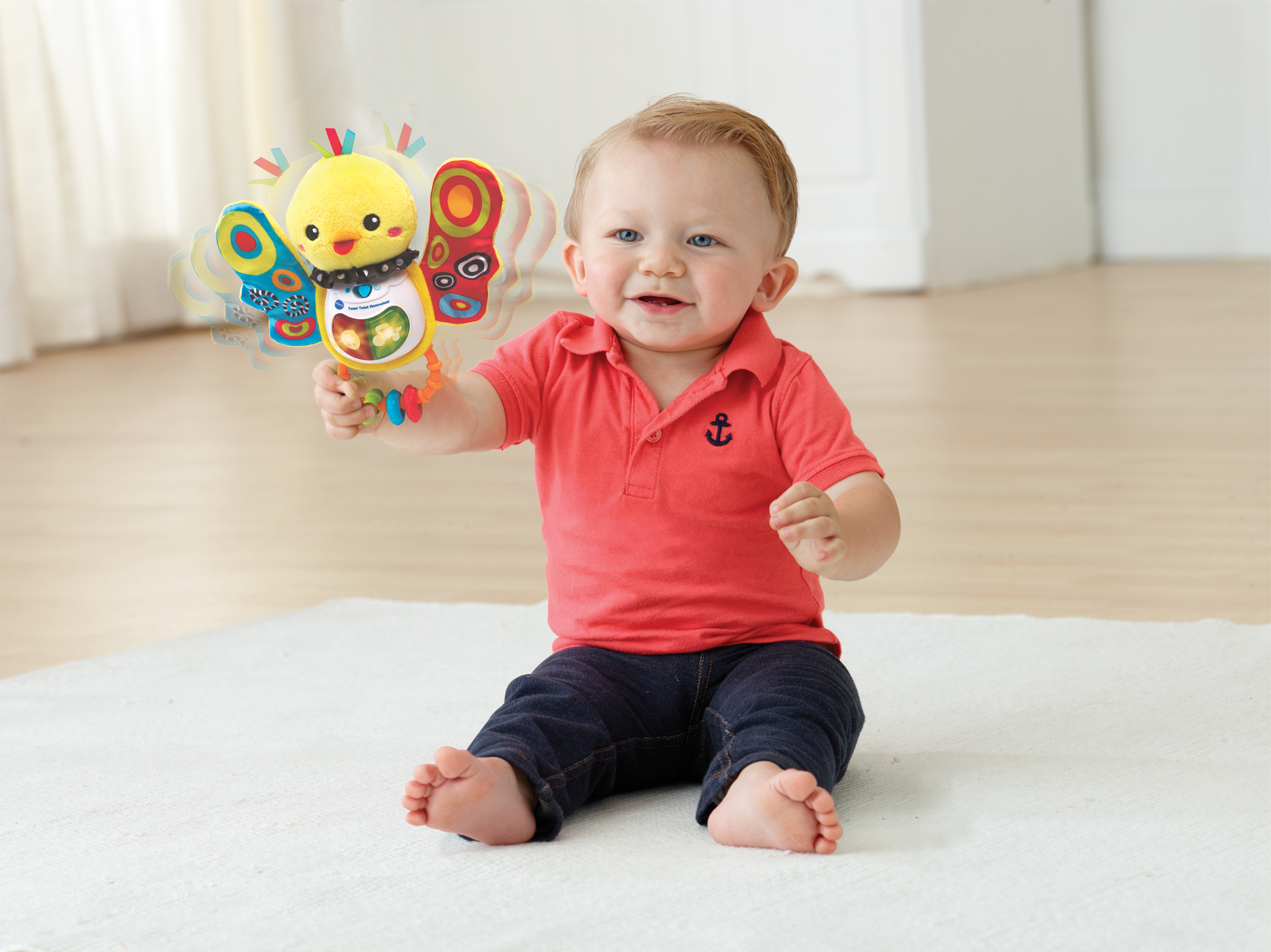 Niemowlę trzymające zabawkę motyla, siedzące na białym dywanie, uśmiechnięte. Czerwona koszula i niebieskie dżinsy.