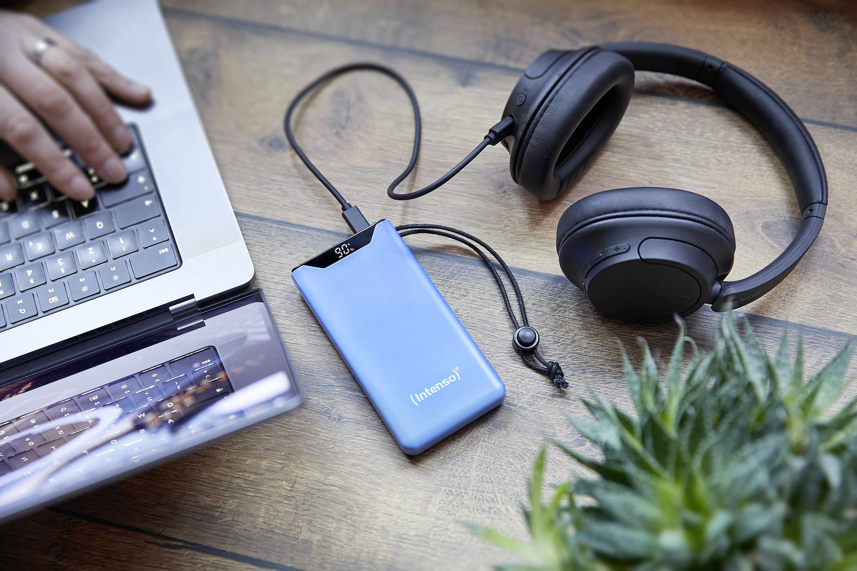 Een laptop, koptelefoon en een powerbank met een display, samen met een kleine plant, op een houten oppervlak.