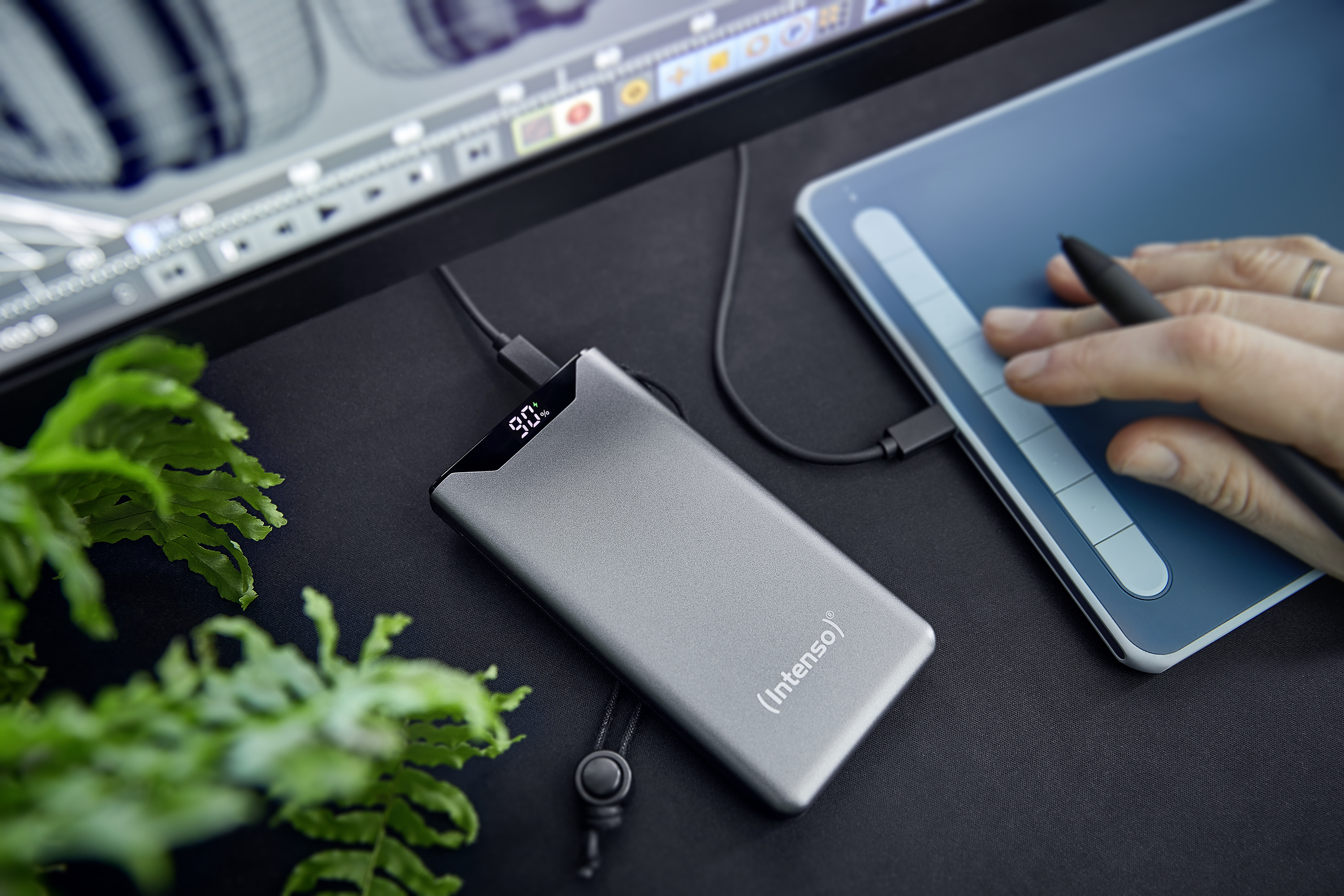 Een zilveren powerbank die een tablet oplaadt met een stylus, op een zwart bureau.