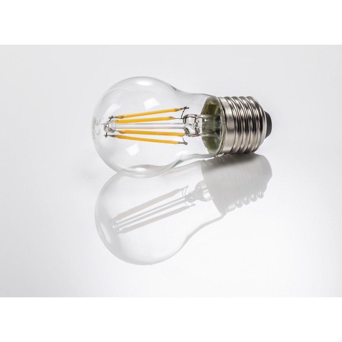 Ampoule transparente avec filaments jaunes, se reflétant sur une surface blanche et grise.