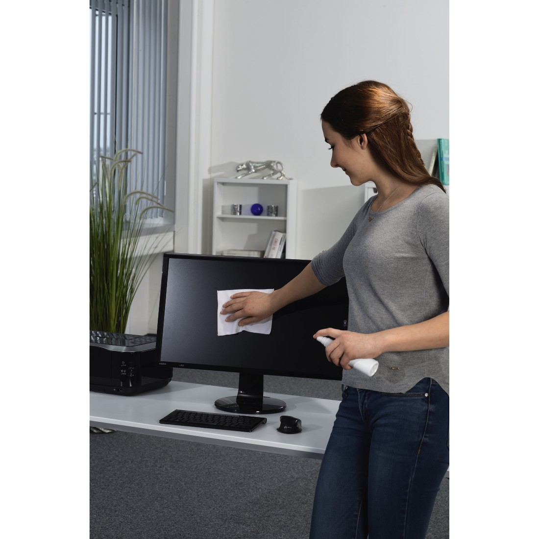 Kobieta czyści monitor komputera na biurku za pomocą butelki z rozpylaczem i szmatki w biurze.