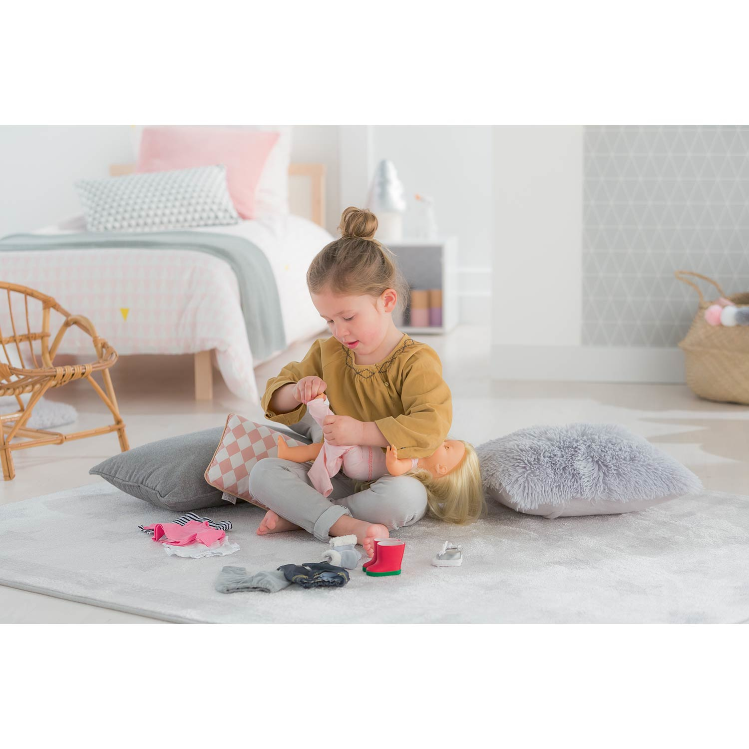 Dziewczynka bawiąca się lalką, zakładająca skarpetki i buty na szarym dywanie.