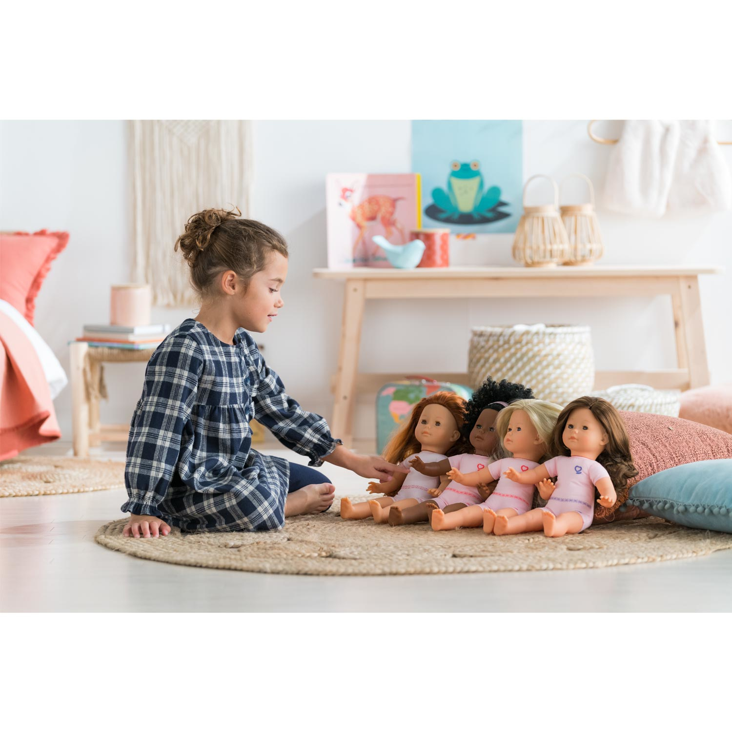 Dziewczynka siedzi z lalkami o różnych odcieniach skóry na dywanie, otoczona zabawkami i meblami.