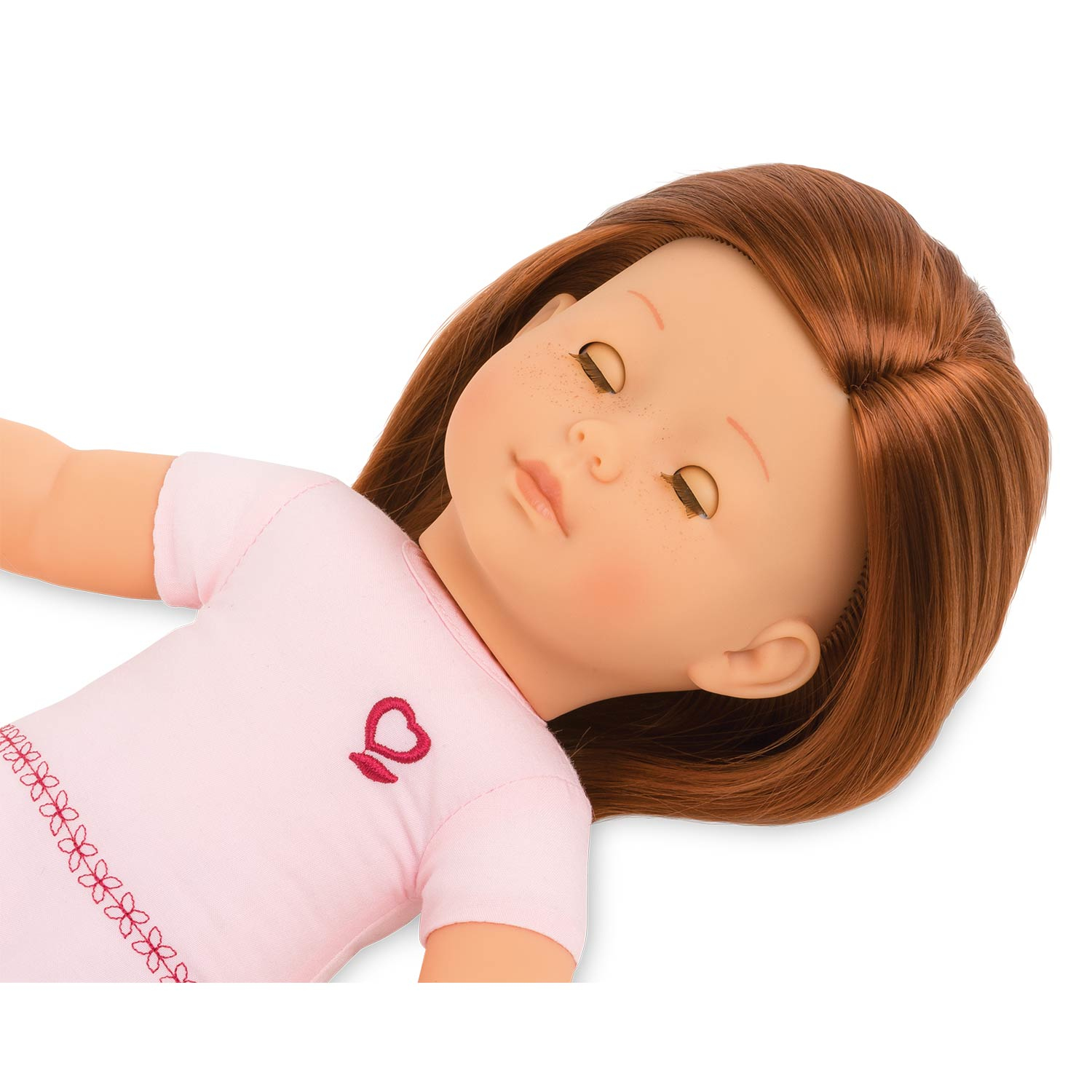 Lalka leżąca. Ma brązowe włosy, różową sukienkę z sercem i piegi na twarzy.