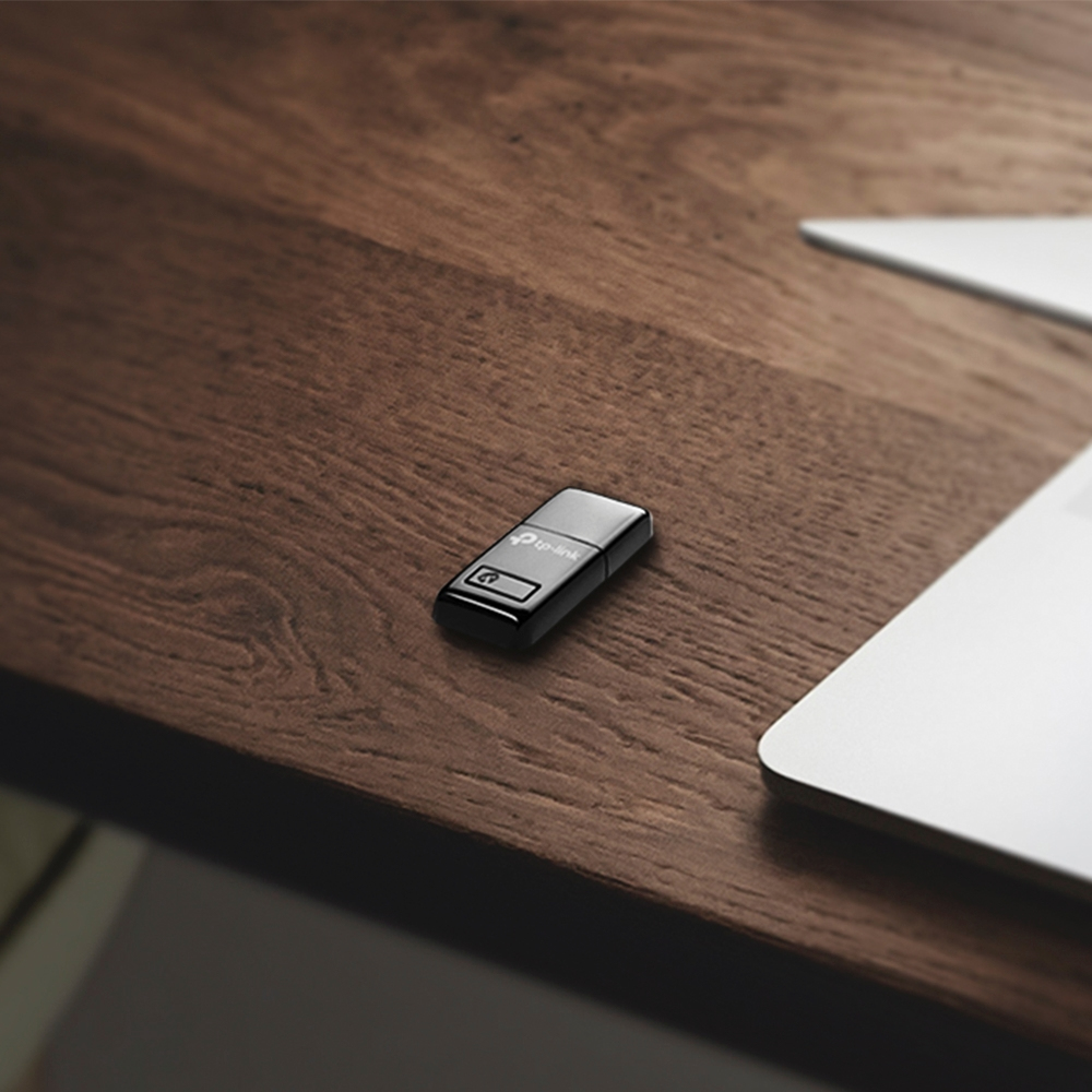 Czarny adapter USB TP-Link na drewnianym biurku, laptop w tle.