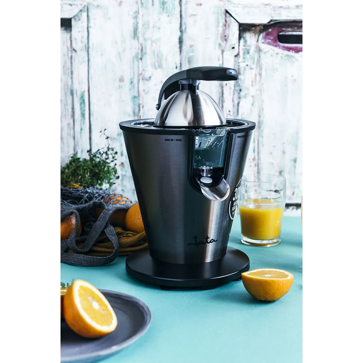 Una exprimidora de cítricos de acero inoxidable con un vaso de zumo y rodajas de naranja sobre una superficie azul.
