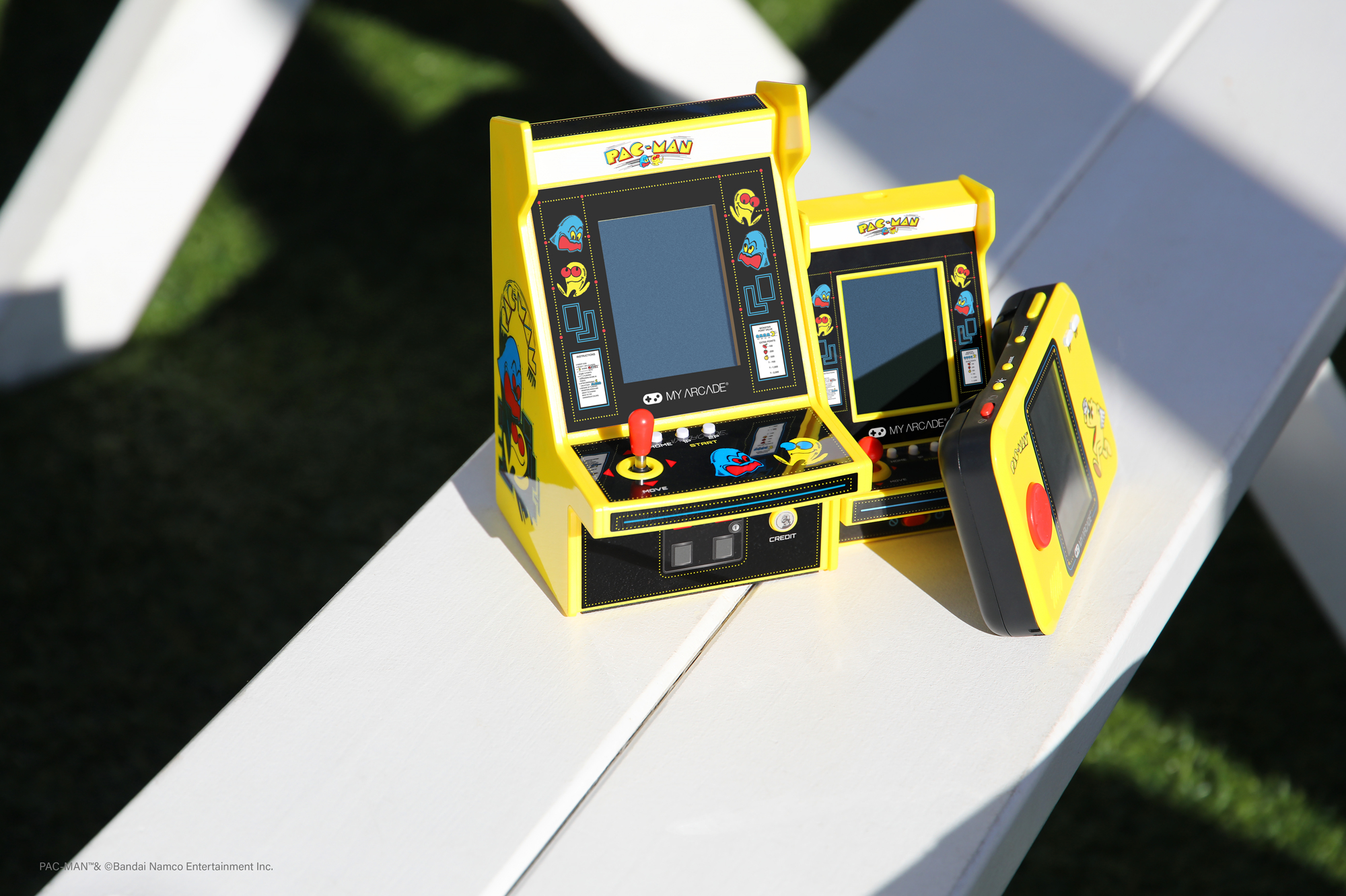 Trzy żółte automaty do gier Pac-Man na białej ławce. Jeden to wersja przenośna.