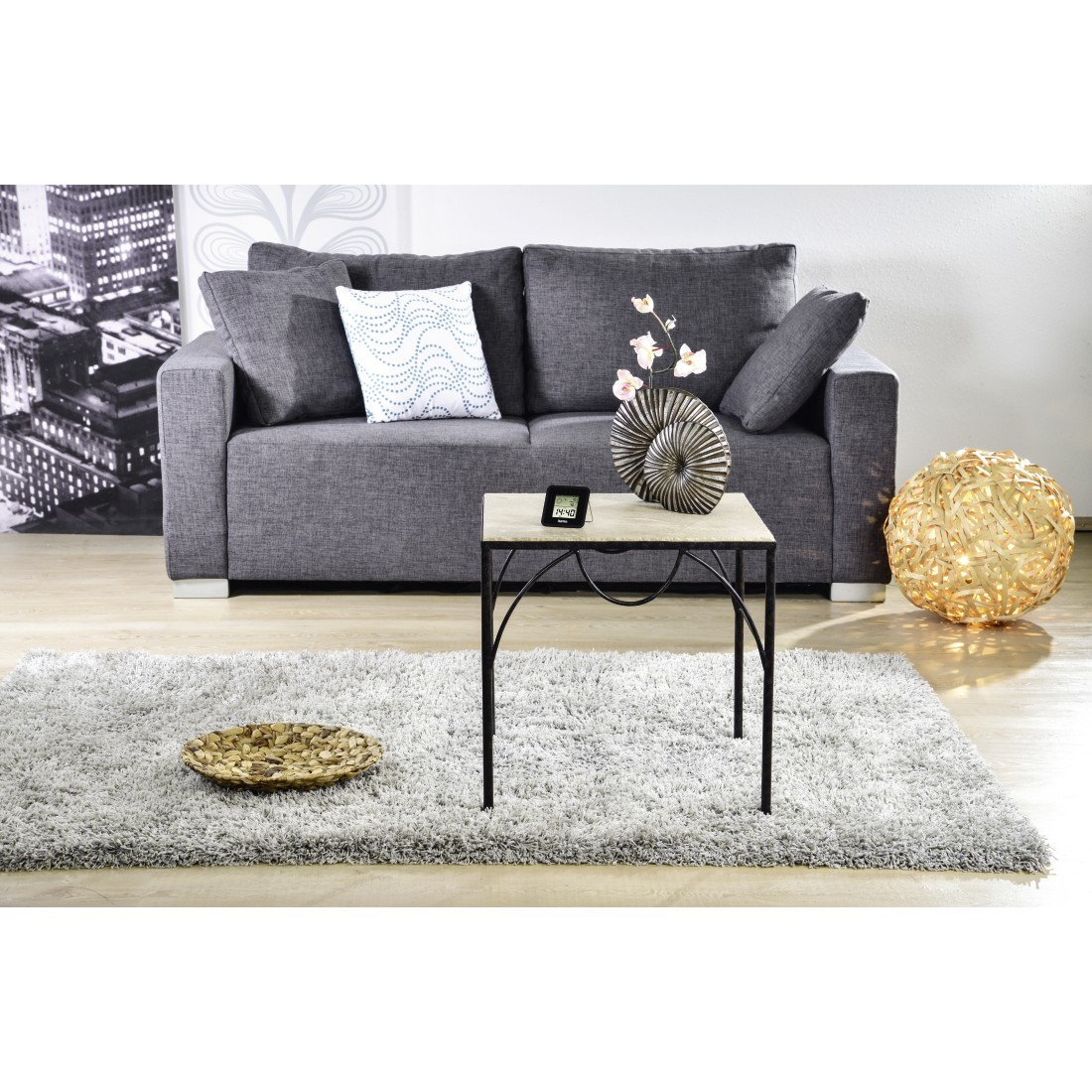 Szara sofa, stół i dywan w pokoju. Obecne są również kwiaty i dekoracyjna kula.