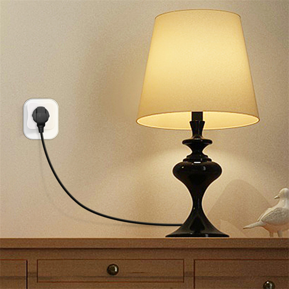 Eine Lampe mit beigem Lampenschirm steht auf einem Holztisch mit eingestecktem Netzkabel. Eine weiße Vogelfigur ist sichtbar.