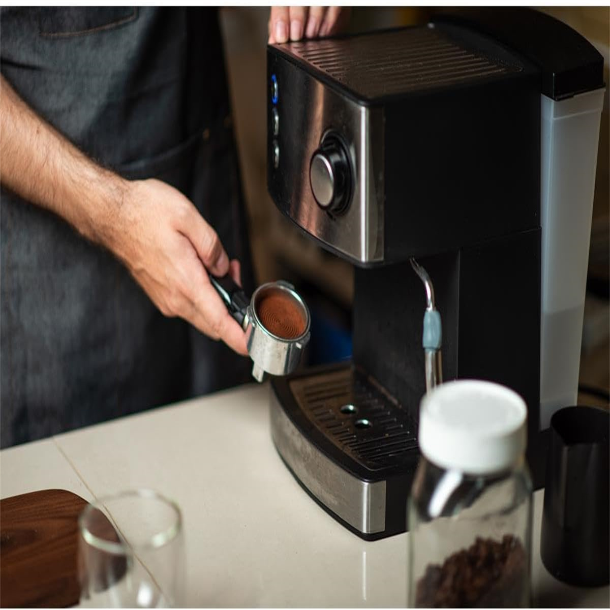 Eine Person bereitet Kaffee mit einer schwarzen Espressomaschine zu. Ein Glas Bohnen und eine Tasse sind sichtbar.