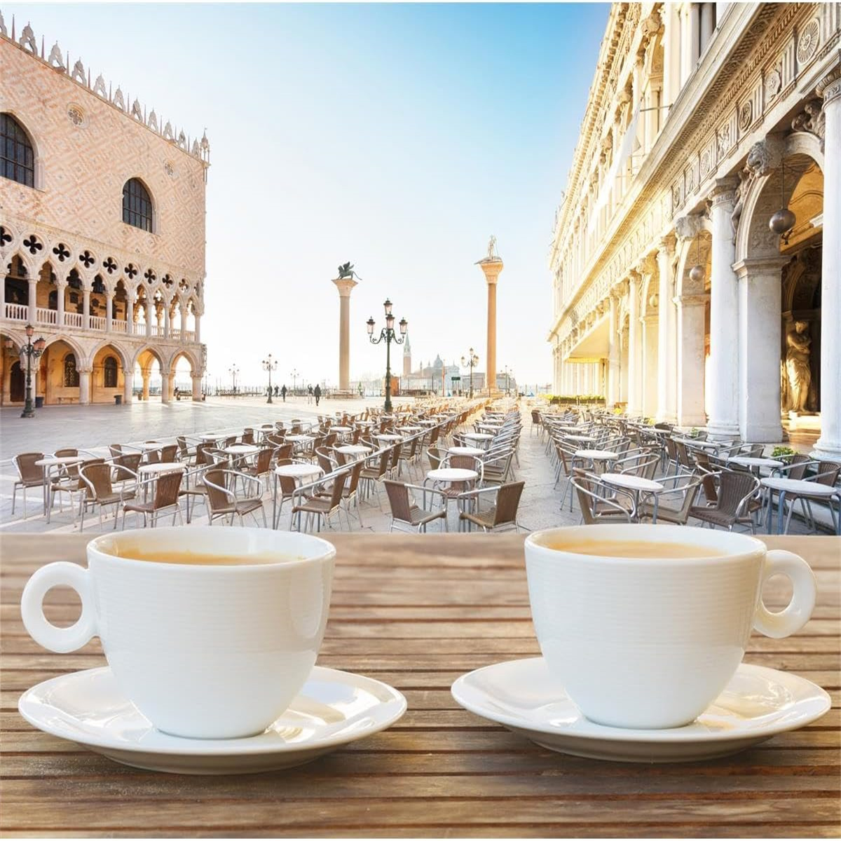 Zwei weiße Kaffeetassen und Untertassen auf einem Holztisch mit Blick auf einen venezianischen Platz mit Gebäuden.