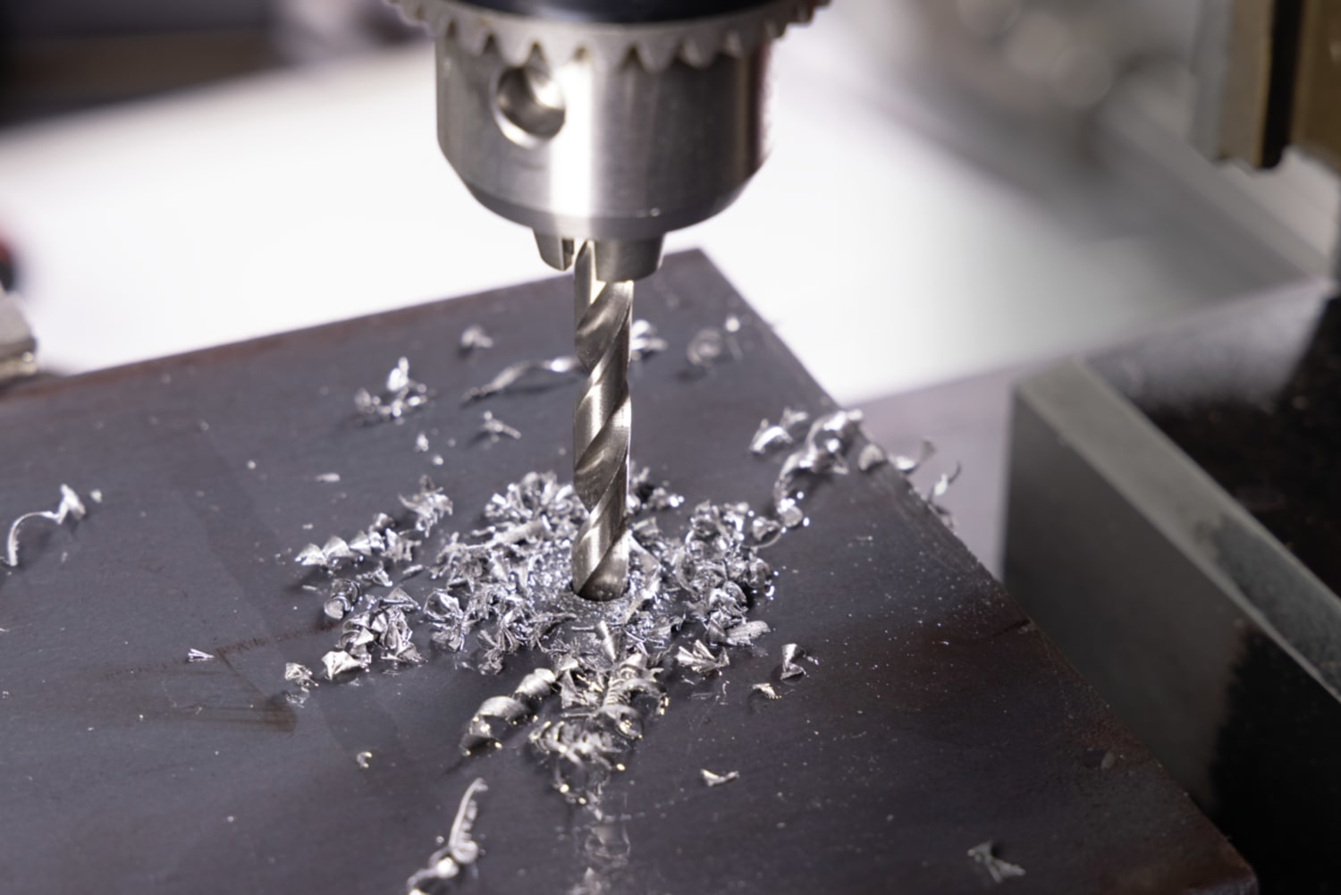 Zbliżenie wiertła wiercącego otwór w metalowej płycie, widoczne wióry metalowe.