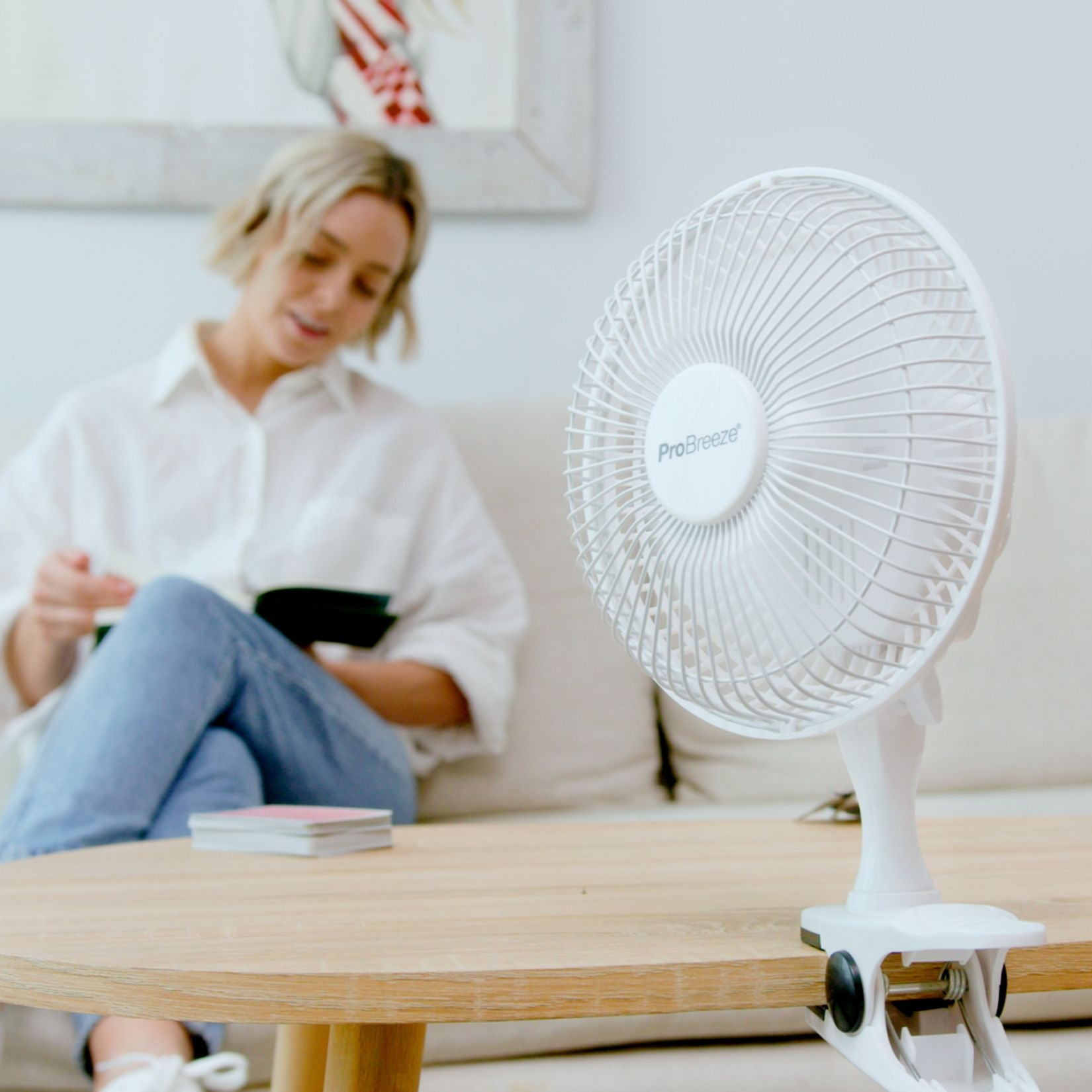 Een witte ventilator op een houten tafel. Een vrouw leest een boek op de achtergrond.
