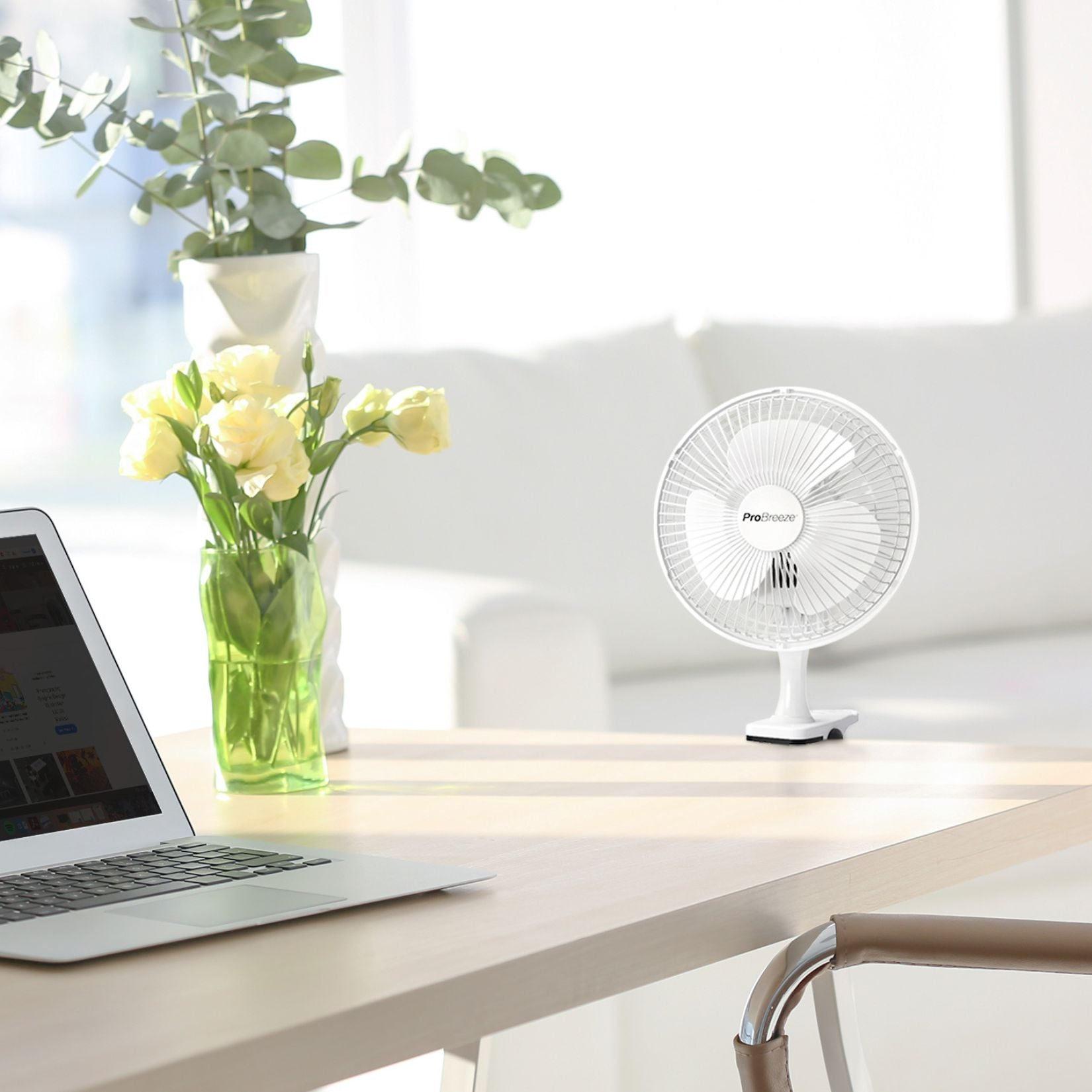 Witte ventilator op houten tafel, laptop en bloemen in vazen, lichte achtergrond.