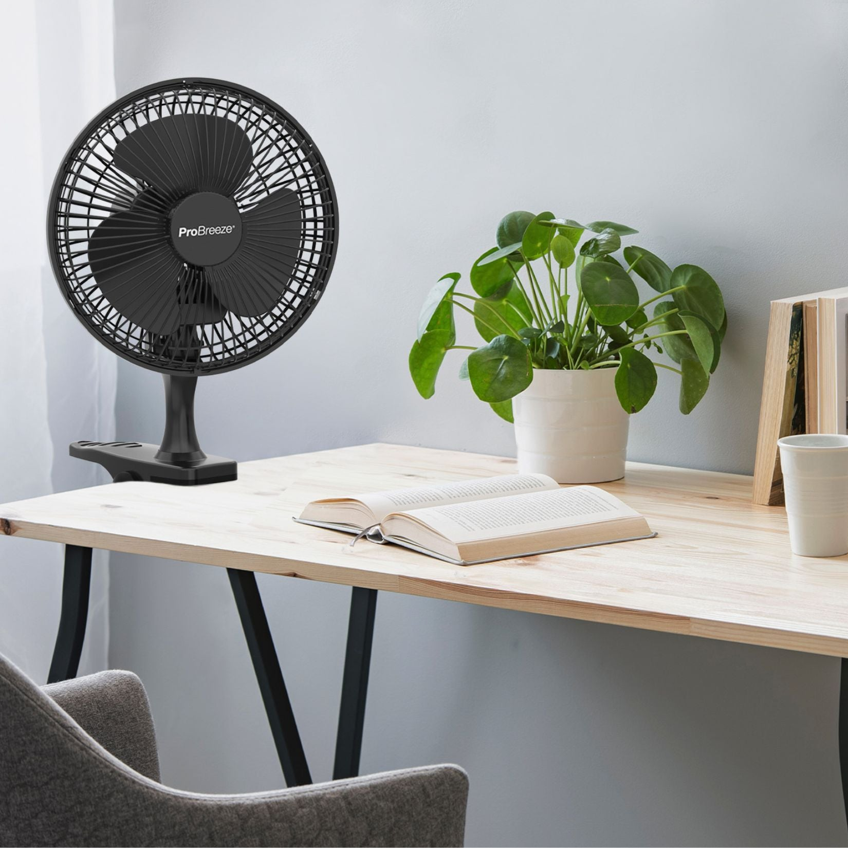 Un ventilatore nero su un tavolo accanto a un libro, una pianta e una tazza, con una sedia grigia in primo piano.