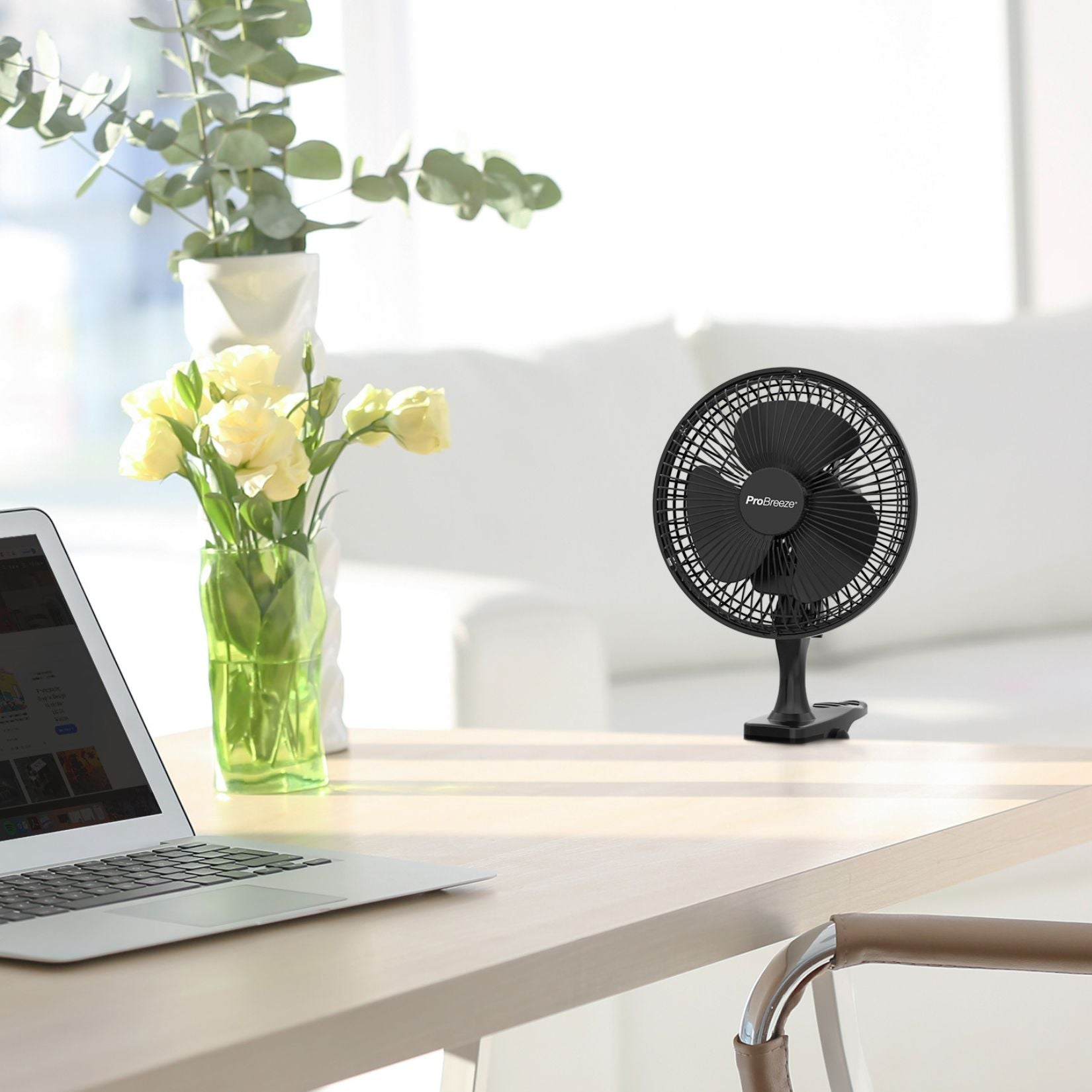 Ventilatore nero su un tavolo, laptop e fiori in vaso. Divano bianco.