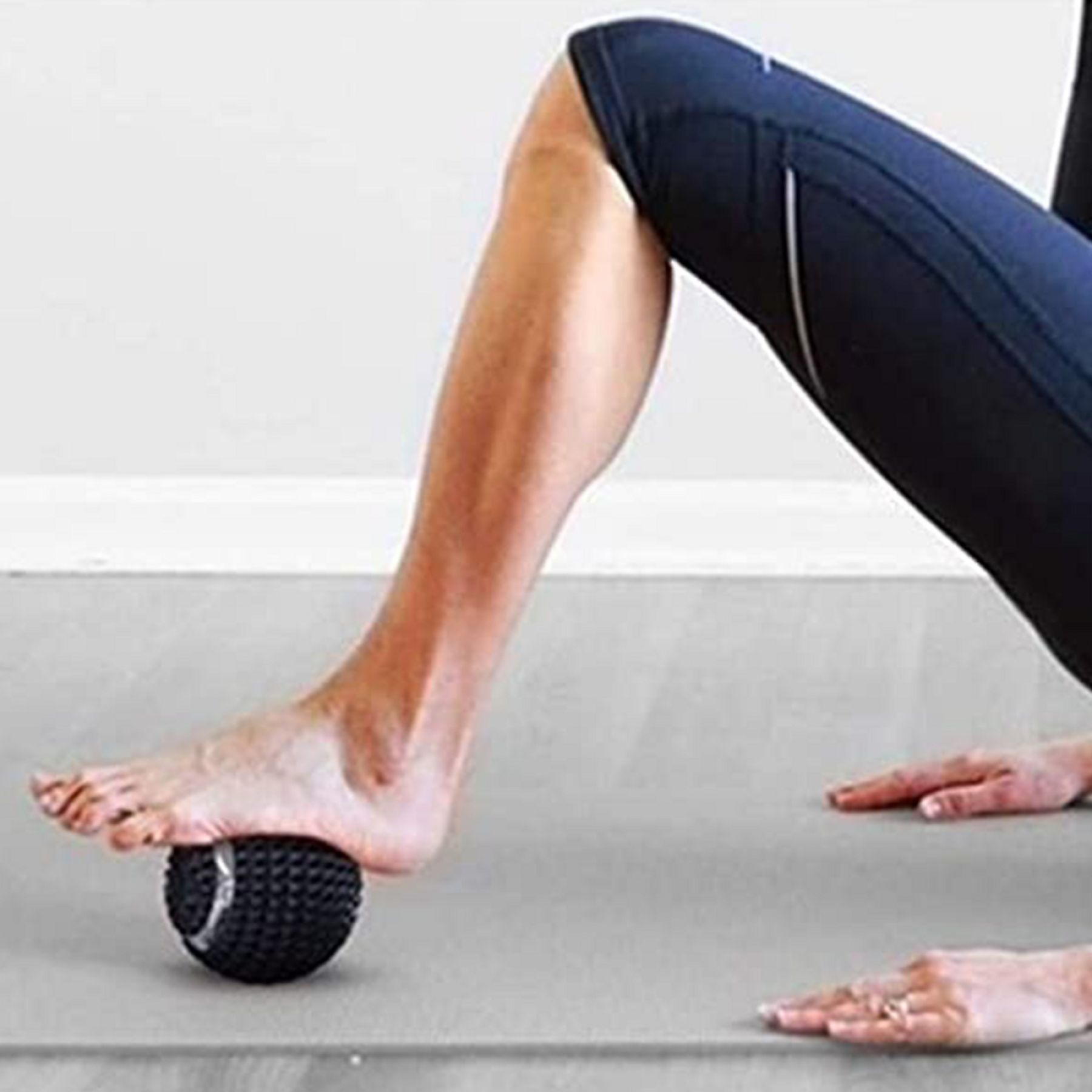 Person drückt Fuß auf einen schwarzen Massageball, wobei dieser auf einer grauen Yogamatte verwendet wird.