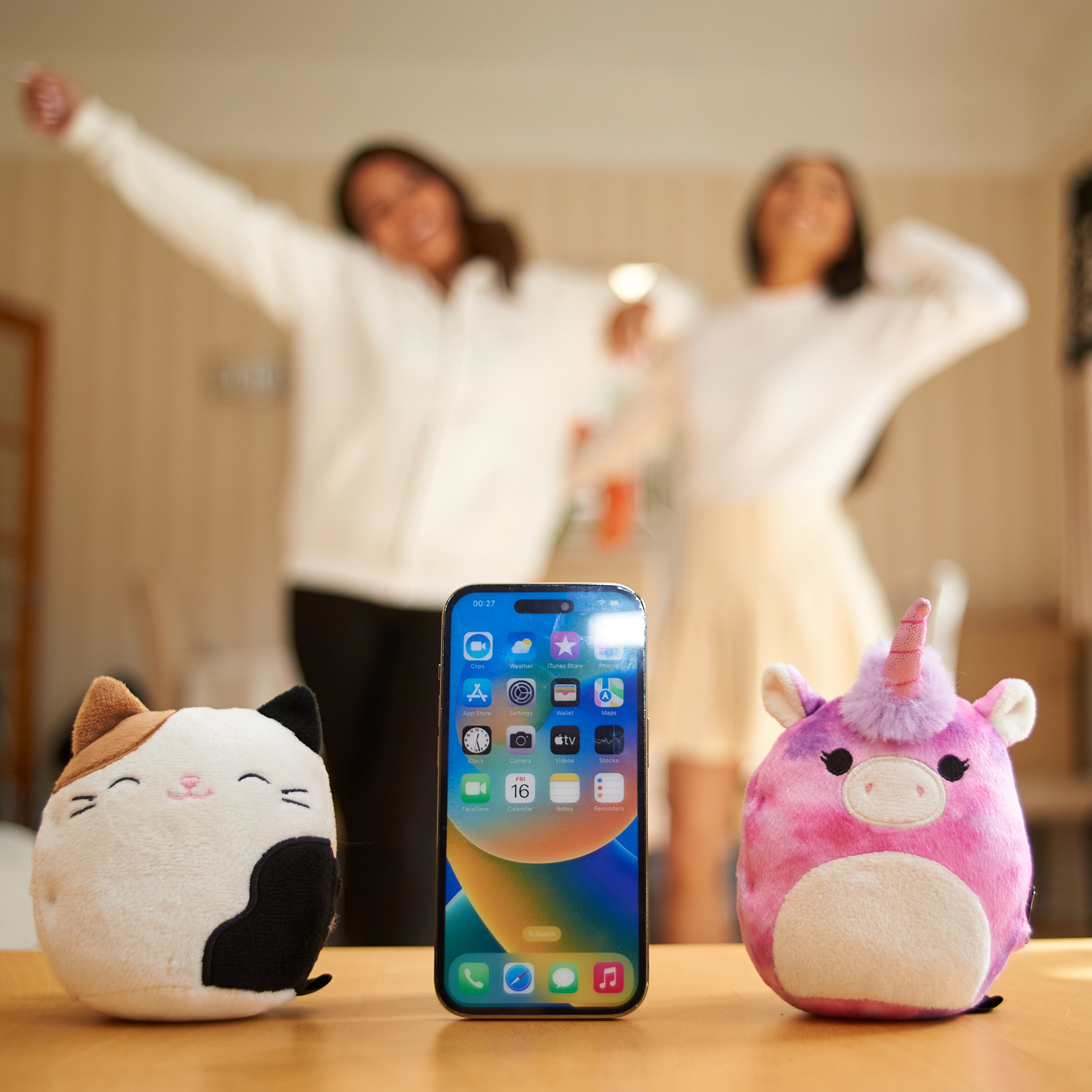 Telefoon tussen knuffels van een kat en een eenhoorn, met twee mensen vaag op de achtergrond.