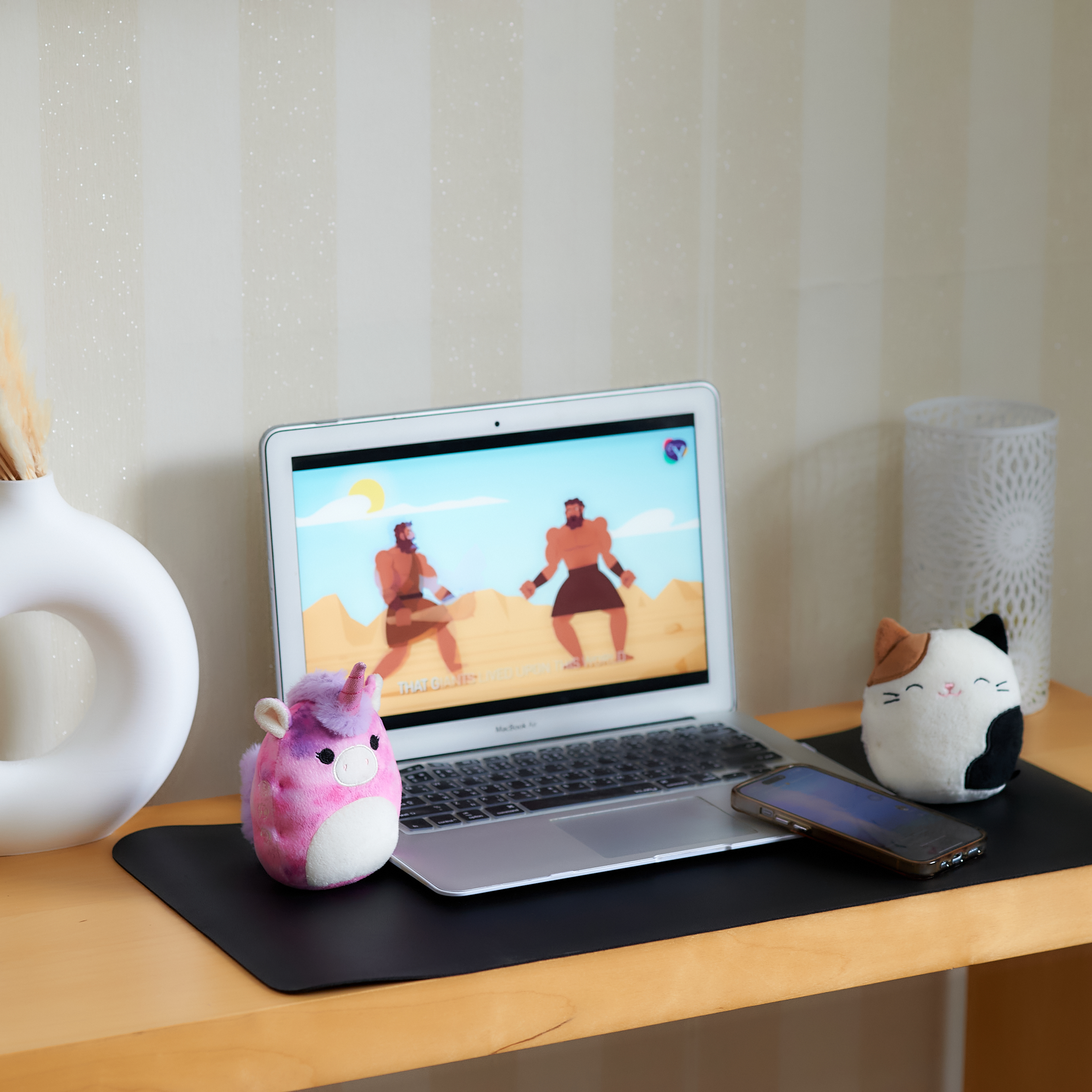 Laptop op een bureau met een video. Een eenhoorn en kat knuffel ernaast. Vaas en lamp op de achtergrond.
