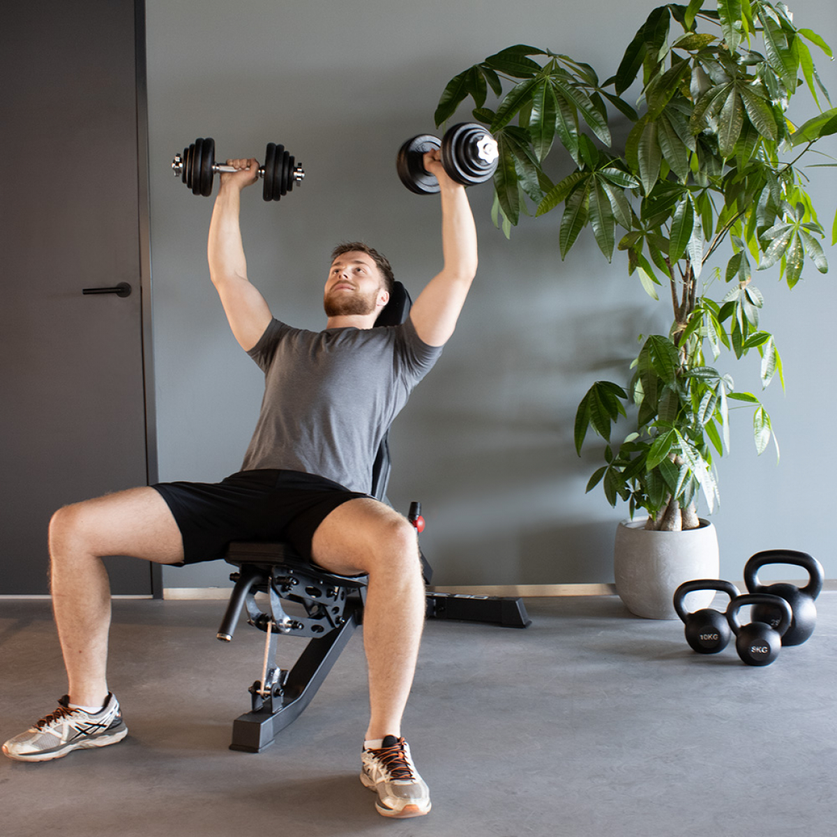 Man lifting weights in a gym. He is seated on a bench.  Dumbbells in hand, plant in the background.