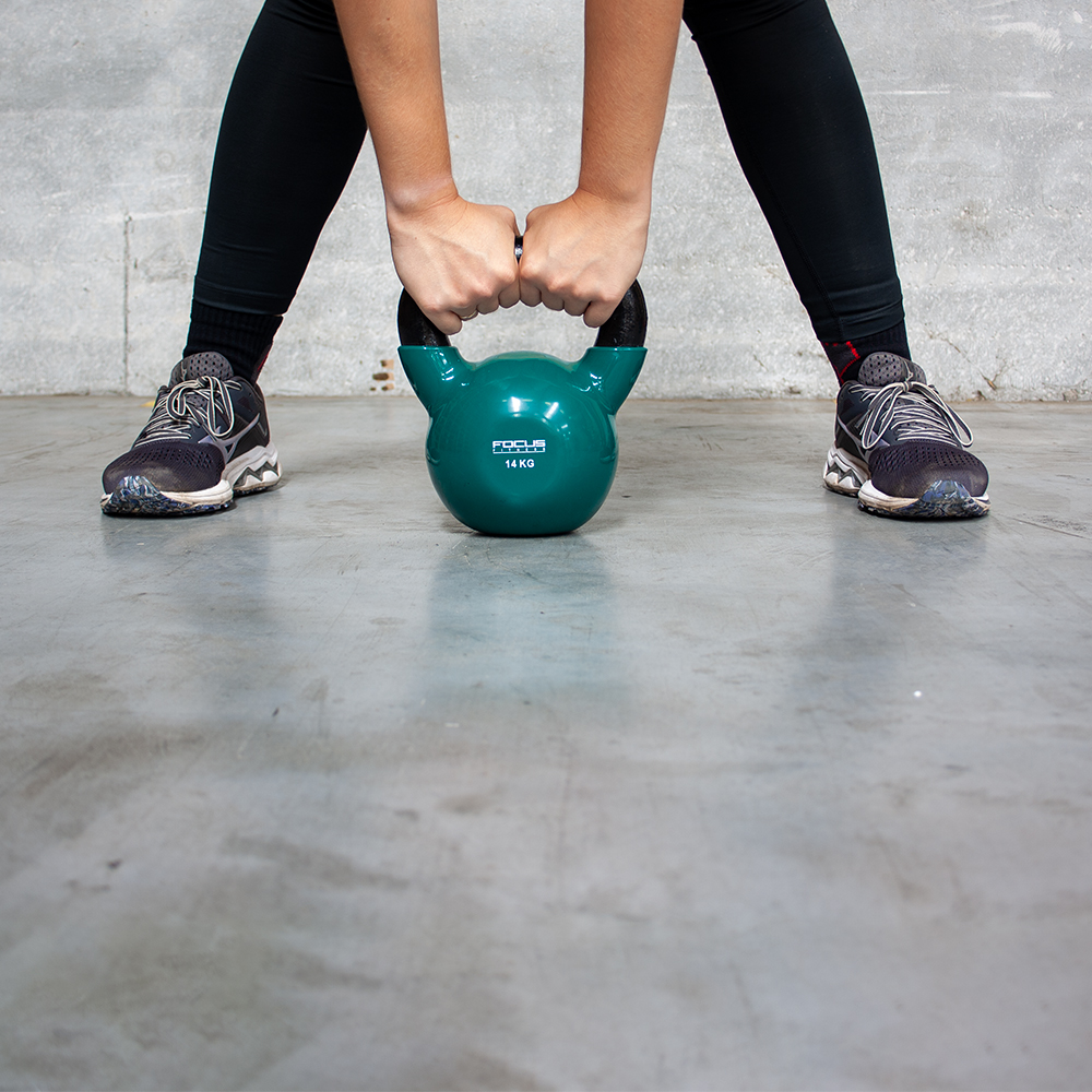 Persoon houdt groene kettlebell vast, benen gespreid, draagt sneakers.