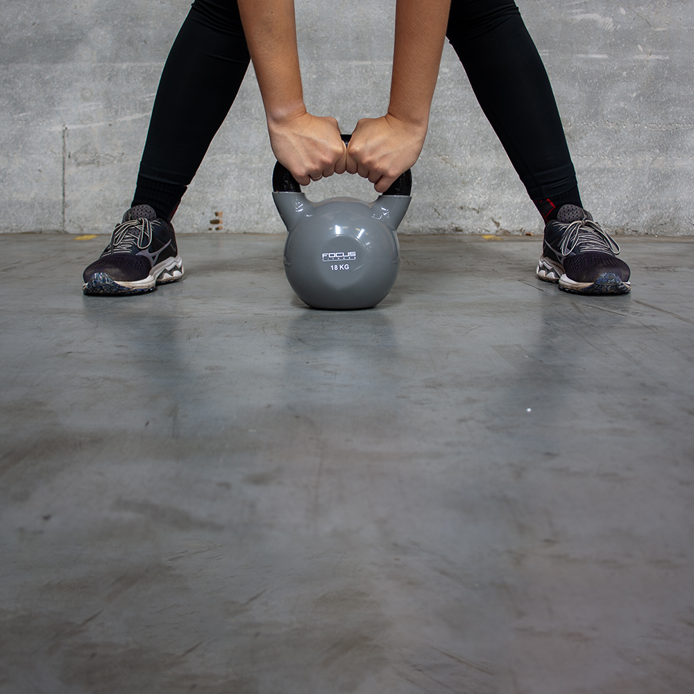 Persoon houdt grijze kettlebell tussen benen op grijze betonnen vloer.