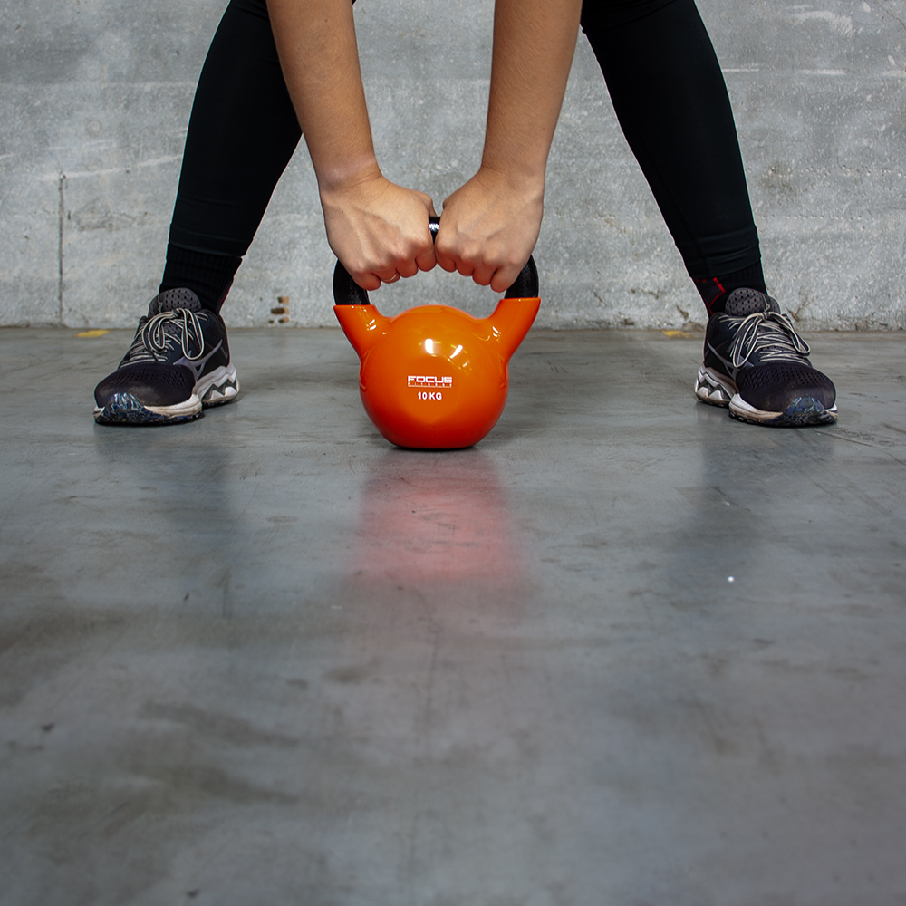 Persoon houdt een oranje kettlebell vast. Benen gespreid, draagt zwarte leggings en schoenen.