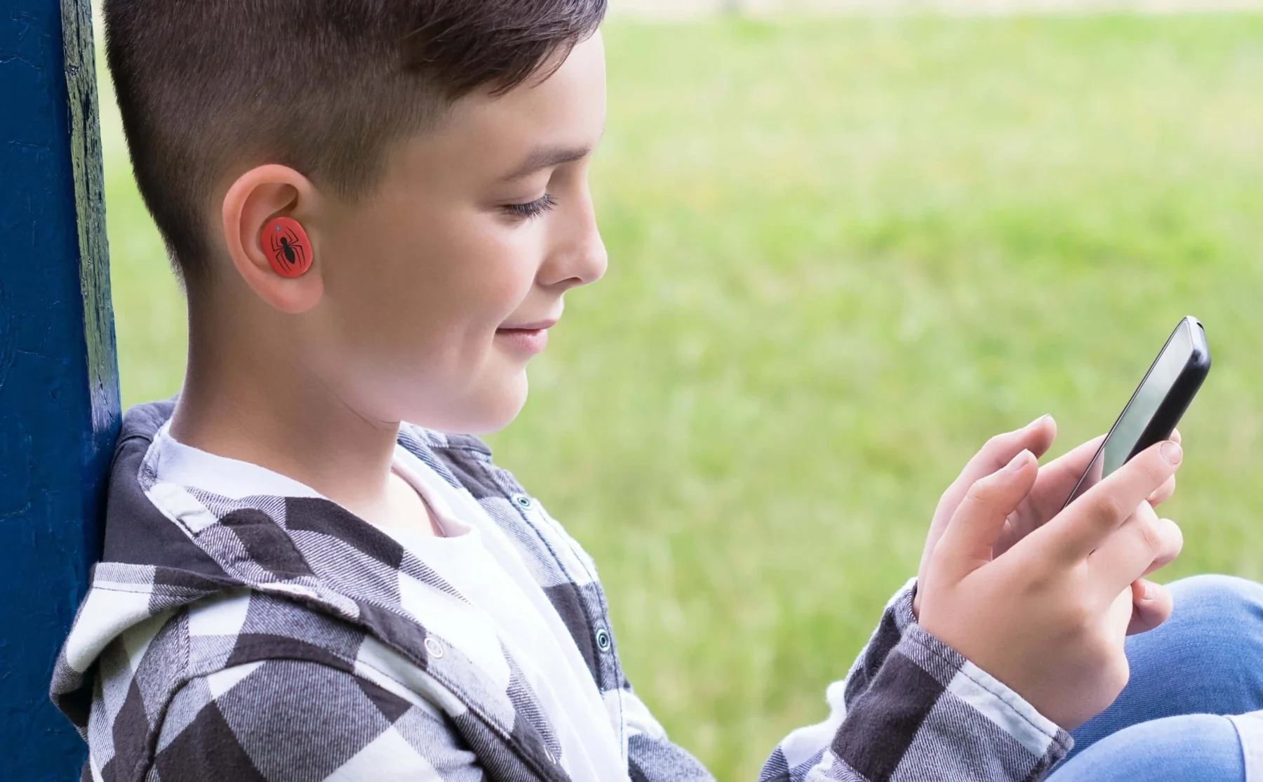 Een jongen met oordopjes kijkt naar een telefoon. Hij draagt een geruit overhemd.