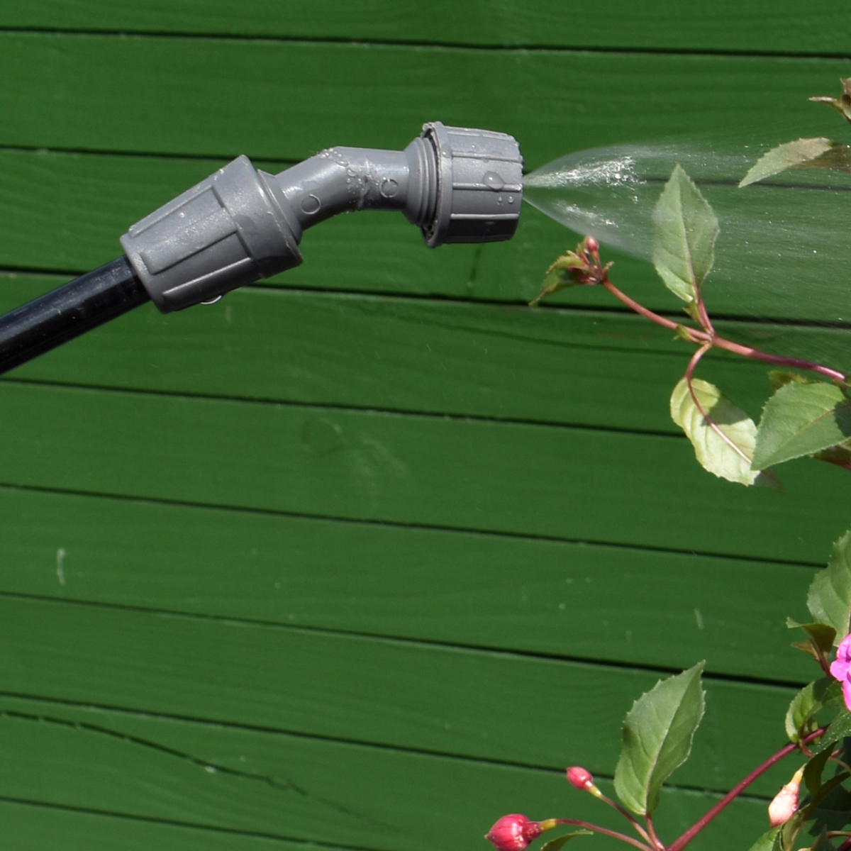 Water sproeit uit een grijs mondstuk. Groene houten schutting en roze bloemen op de achtergrond.
