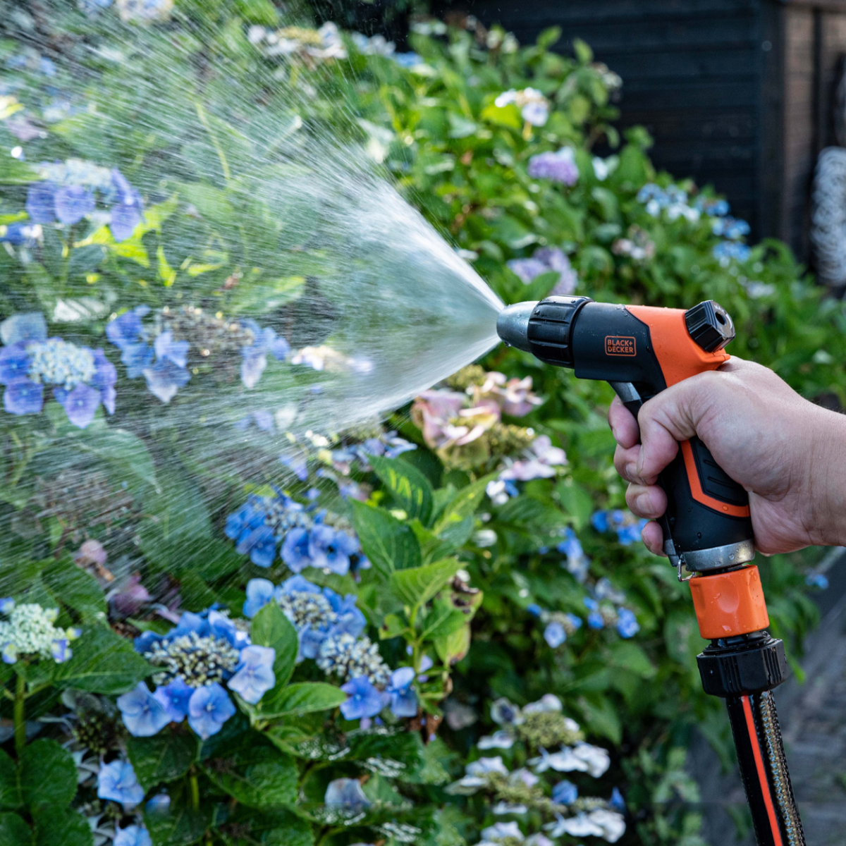 Hand die een zwarte en oranje watersproeier vasthoudt, water sproeit op blauwe bloemen.