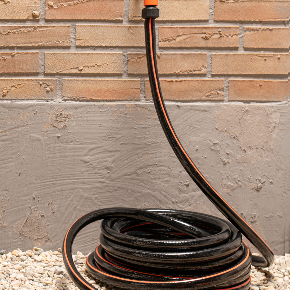 Black garden hose coiled on pebbles, near a brick wall and gray wall.