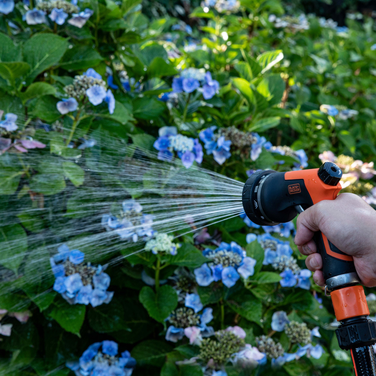 Een hand spuit water uit een zwart-oranje spuitmond op blauwe bloemen.