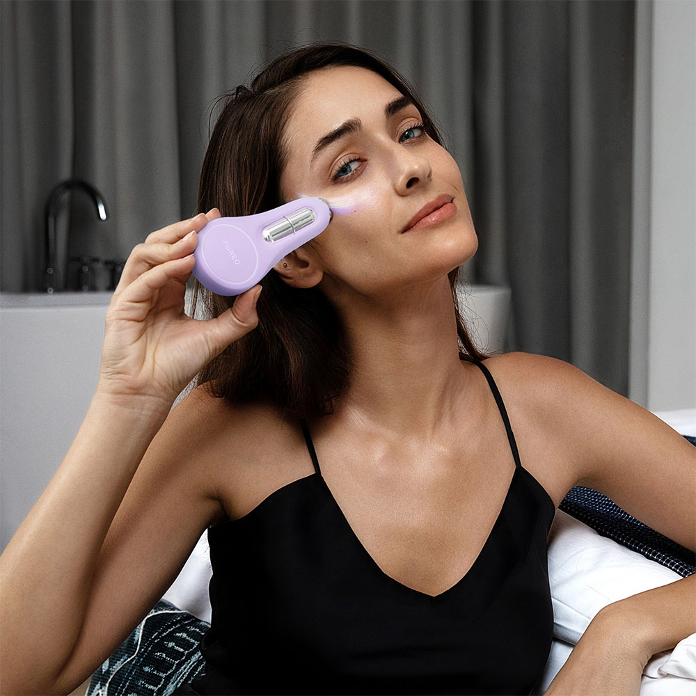 A woman using a purple FOREO device on her cheek, looking at the camera.