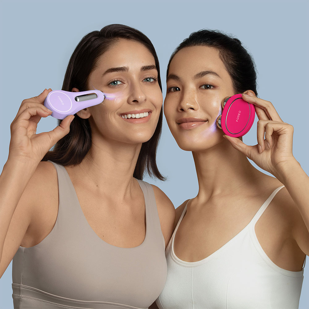 Twee vrouwen die gezichtsbehandelingsapparaten vasthouden, glimlachend. Blauwe achtergrond.