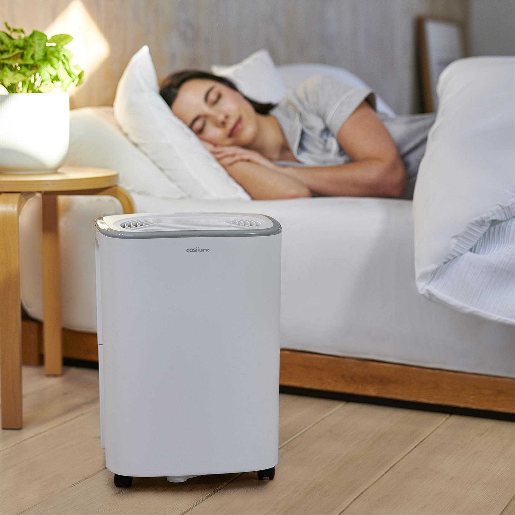 Schlafende Frau im Bett mit einem weißen Luftentfeuchter auf dem Boden. Grüne Pflanze im Hintergrund.