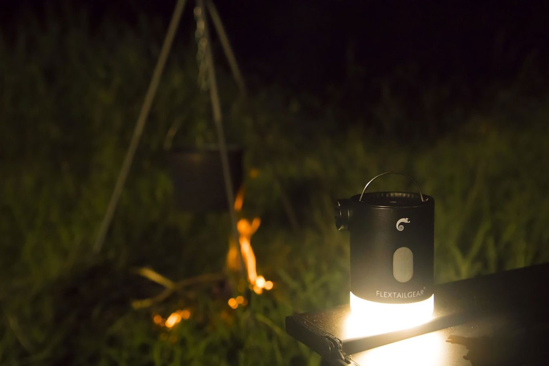 Eine schwarze Laterne auf einem Tisch leuchtet. Ein Lagerfeuer und ein Stativ sind im unscharfen Hintergrund.