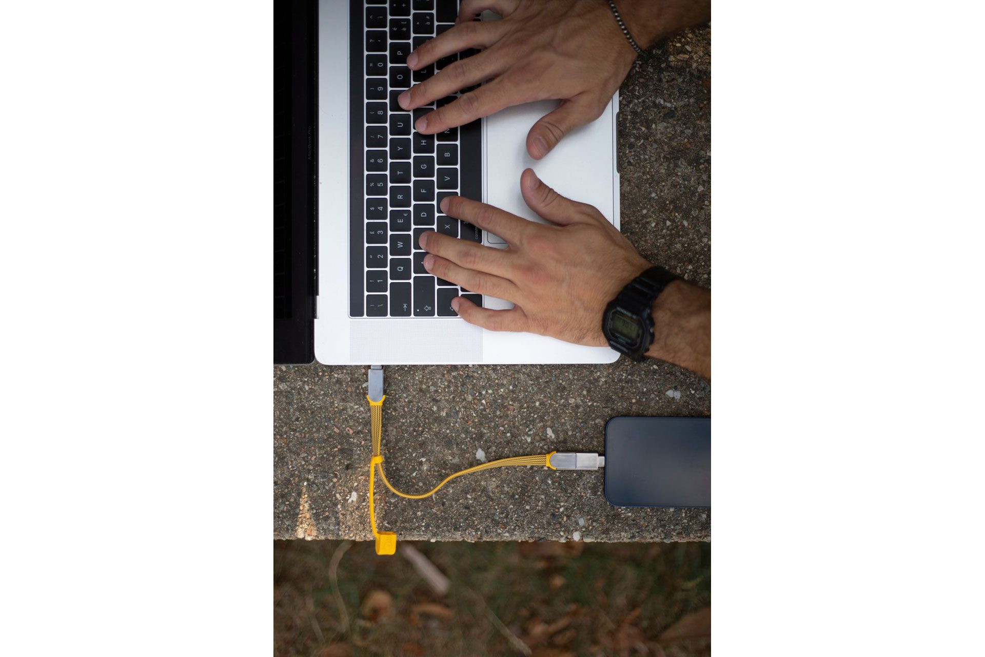 Mains tapant sur un ordinateur portable avec un câble jaune connecté à un téléphone en charge sur une surface en béton.