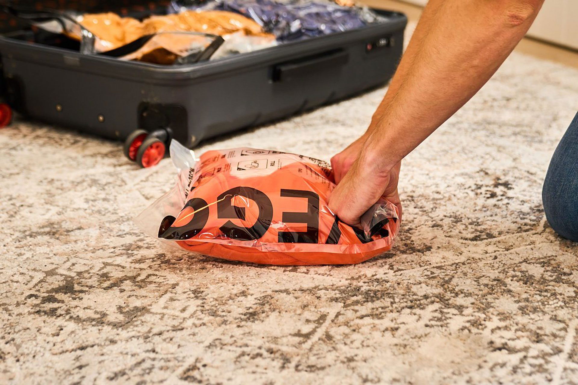 Hand hält ein Paket, ein Koffer im Hintergrund, das Paket ist orange mit weißem Text.