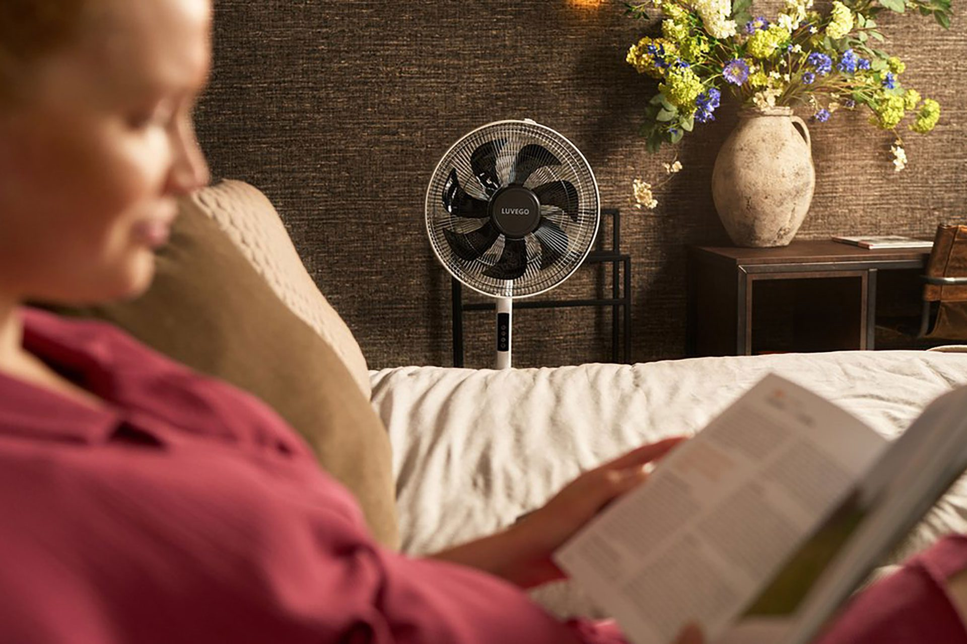 Eine Frau liest auf einem Bett, hinter ihr steht ein weißer Ventilator. Rechts steht eine Blumenvase.